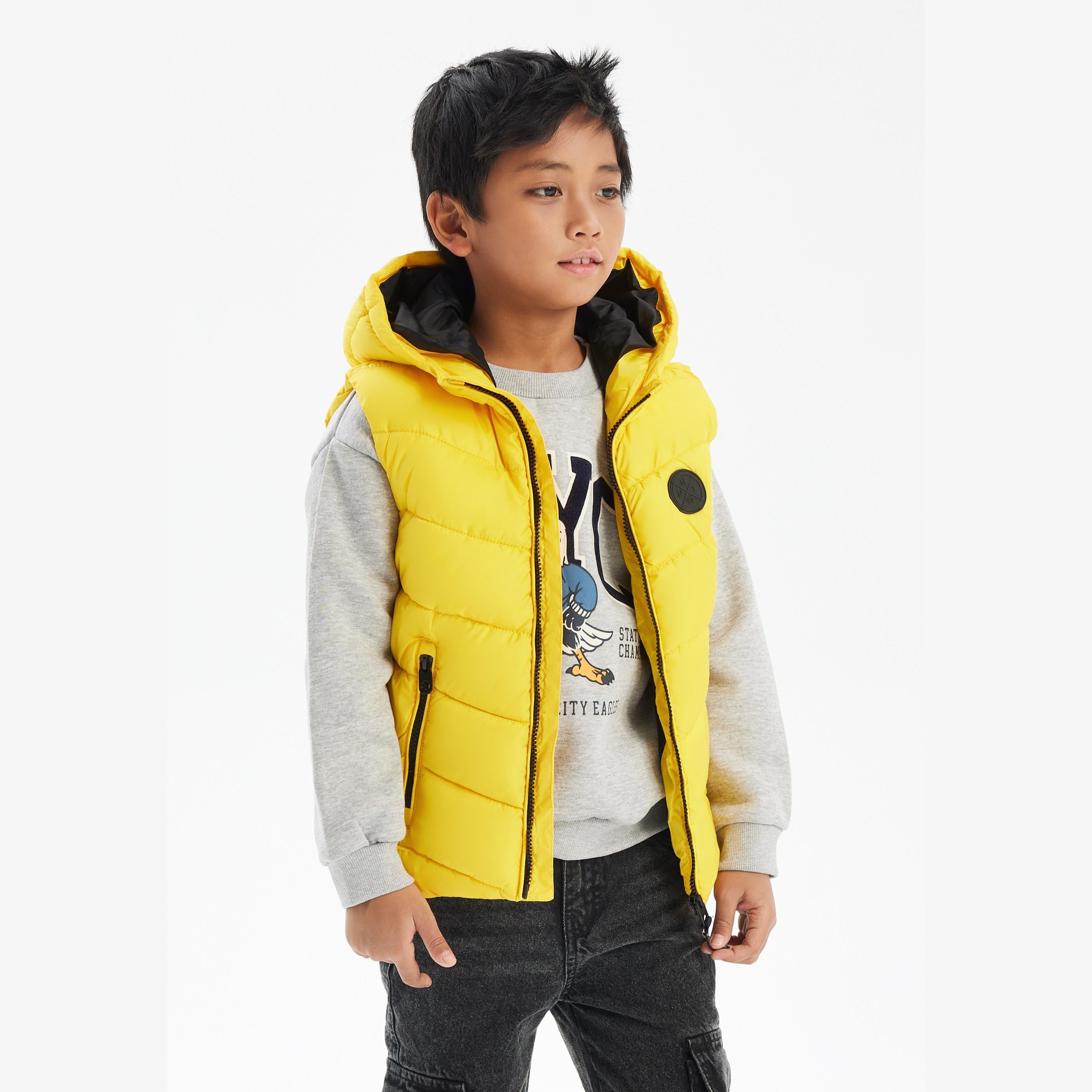 Boy wearing bright yellow padded gilet with black hood over grey sweatshirt and dark jeans against white background
