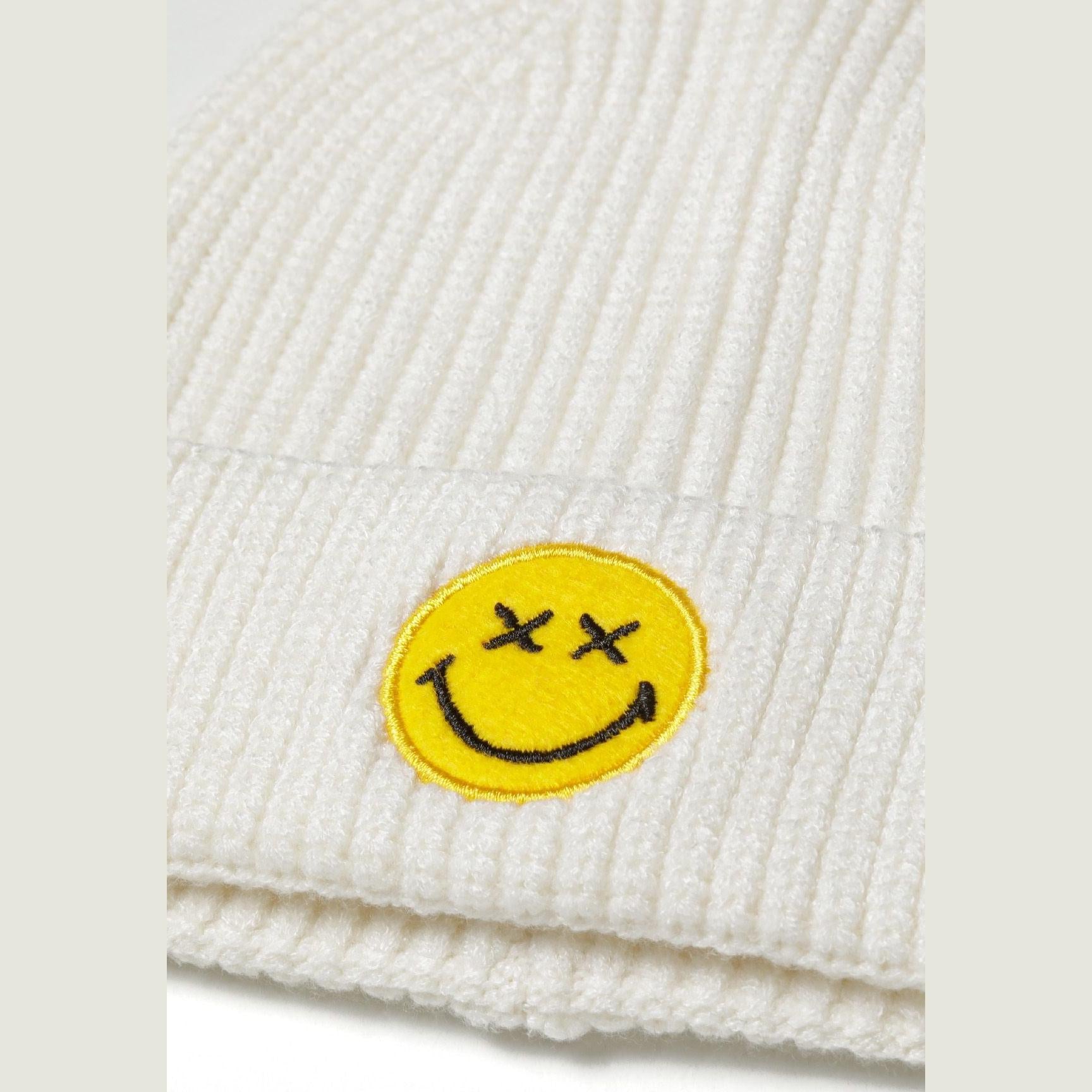 Close-up detail of yellow embroidered smiley face patch with X eyes on cream ribbed beanie fabric