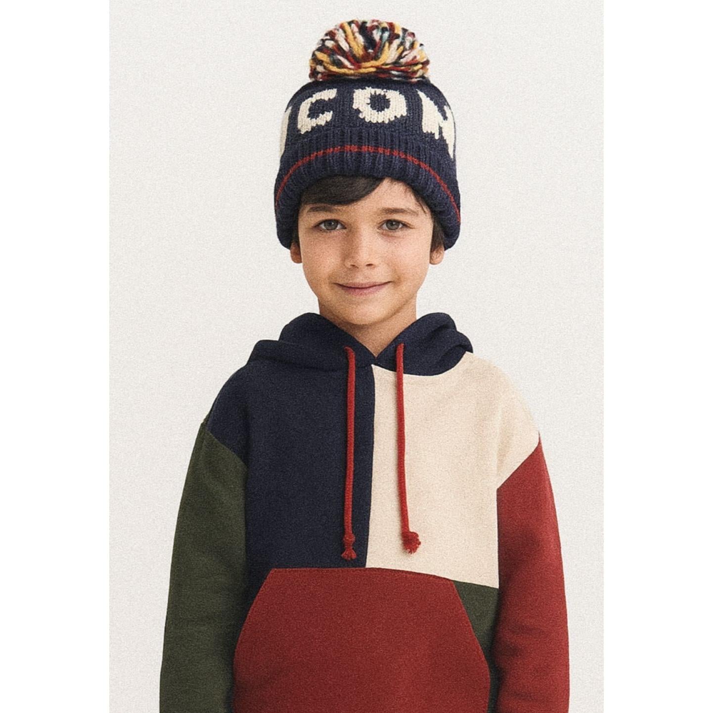 Child wearing navy knitted beanie with white jacquard pattern and multicoloured pom pom, paired with colour-block hoodie against white background