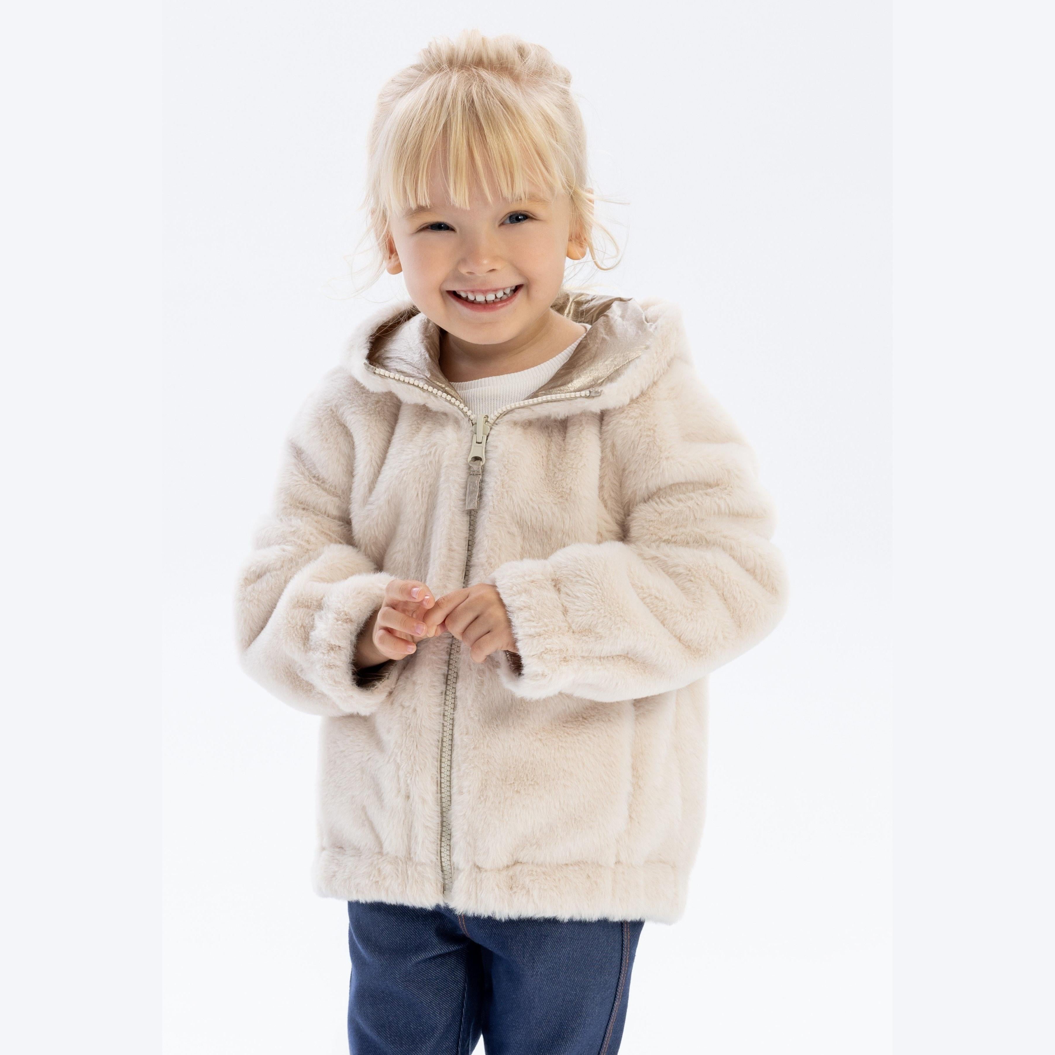 Young girl wearing beige reversible jacket with hood up, smiling at camera against white background, showing metallic exterior side