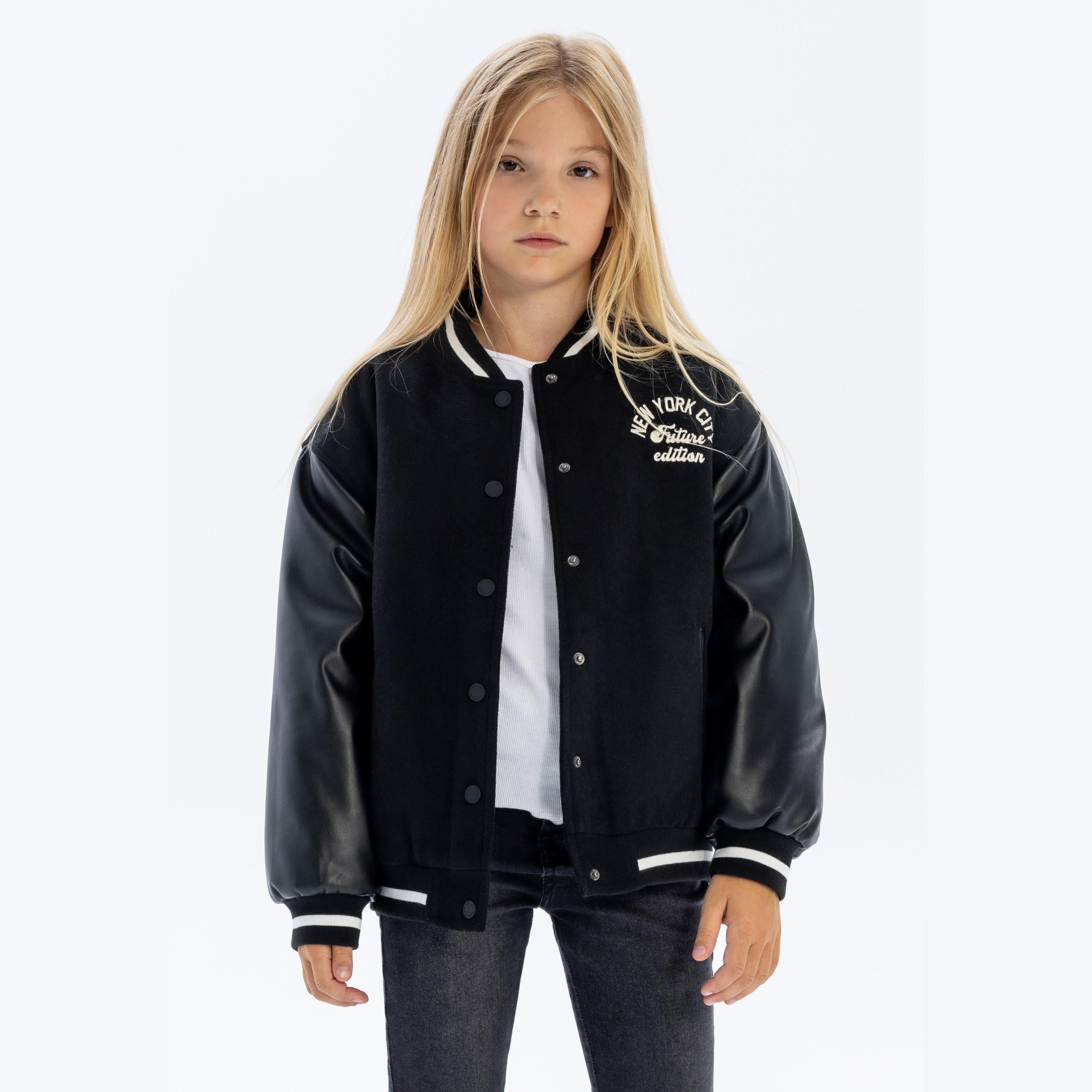 Young blonde girl wearing black baseball jacket with faux leather sleeves over white top and dark jeans, standing against white background
