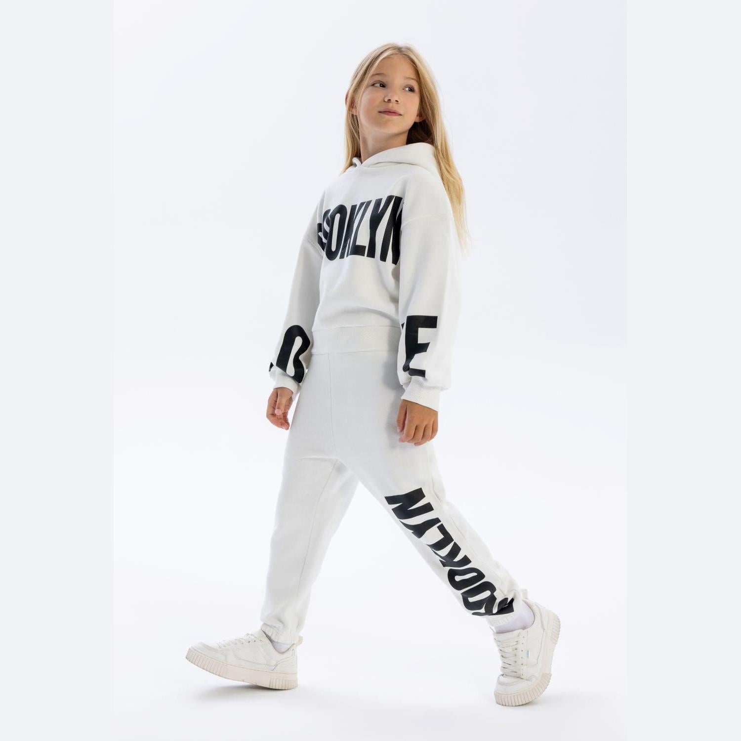 Young girl wearing white hooded tracksuit with black printed text, posed against white background in casual stance