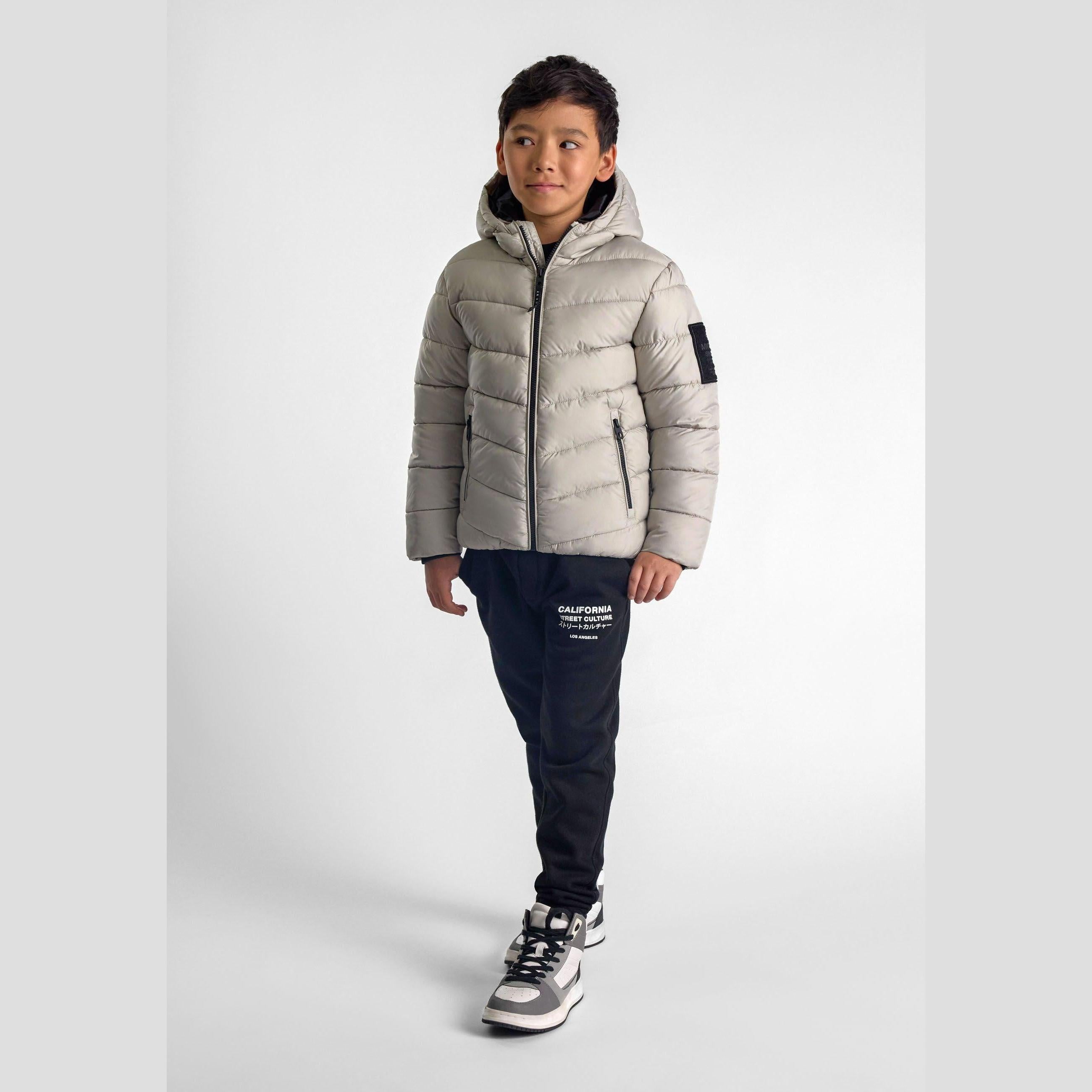 Boy wearing grey quilted jacket with hood and navy joggers against white studio background