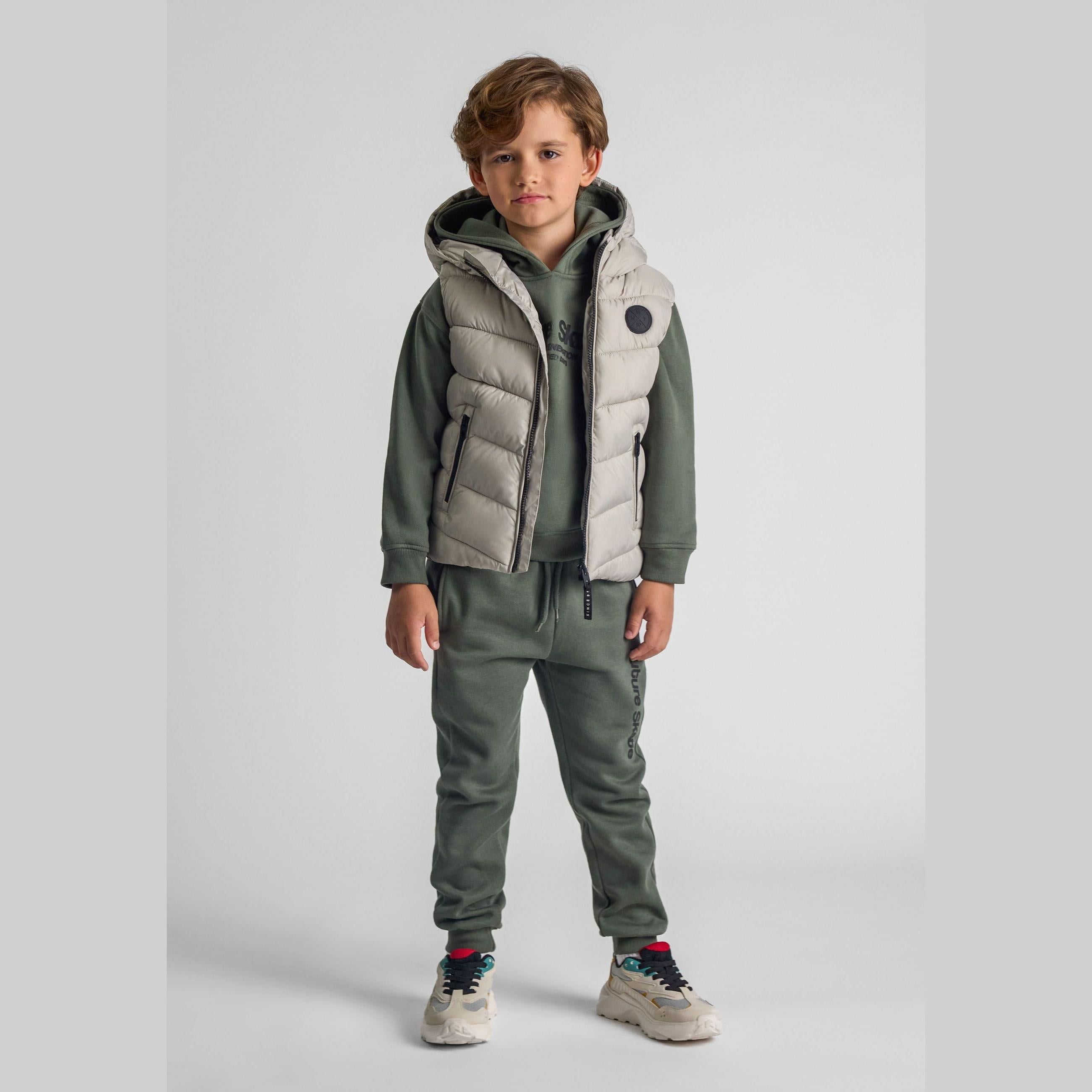 Young boy wearing grey padded gilet with hood over green tracksuit, standing against white studio background
