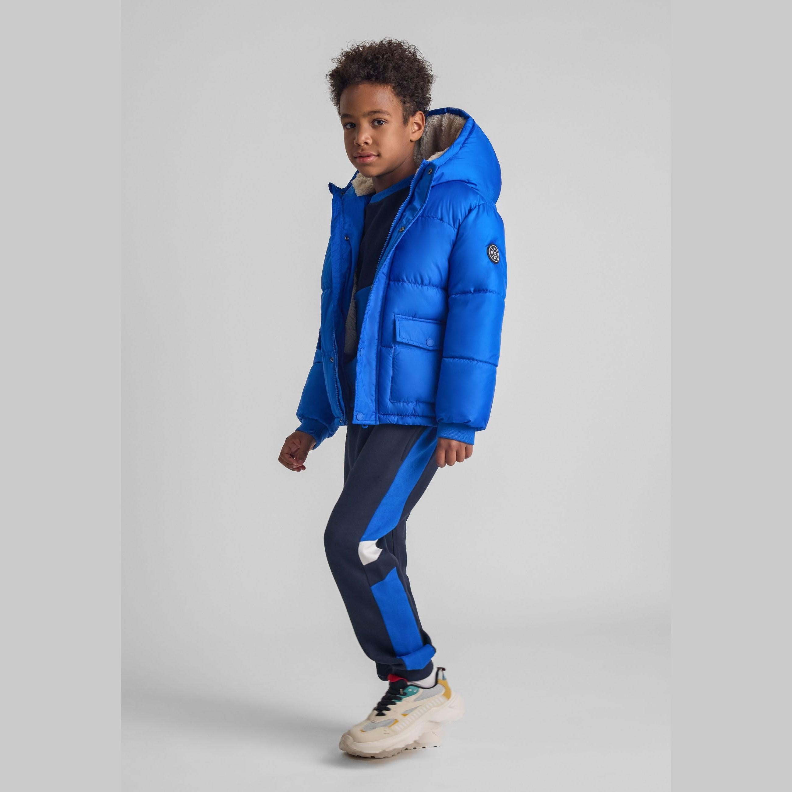 Young boy wearing cobalt blue hooded winter jacket with matching tracksuit bottoms, standing against white studio background