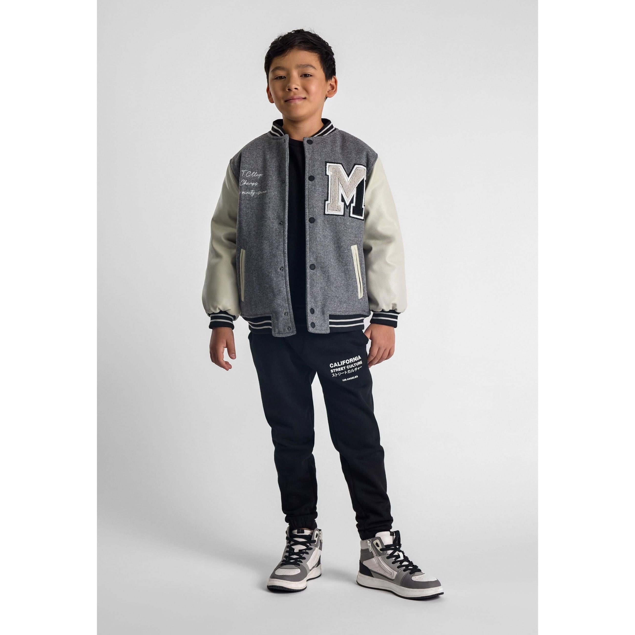 Young boy wearing grey and cream varsity jacket with black trousers, standing against white background