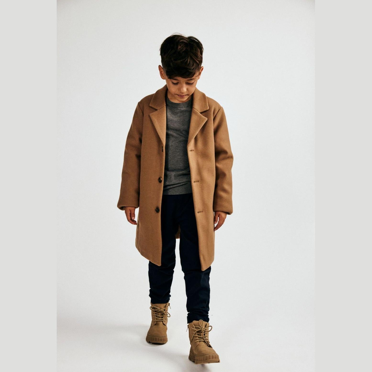 Young boy wearing beige smart coat over grey sweater and dark jeans, styled with brown boots, standing against white background