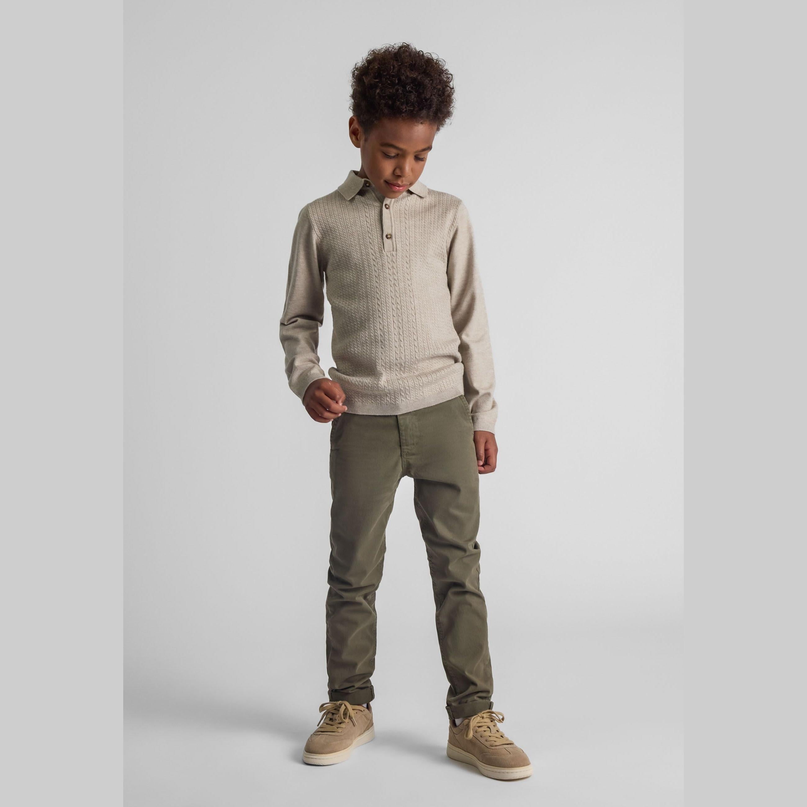 Young boy wearing khaki chino trousers with beige knit polo sweater, standing against white studio background
