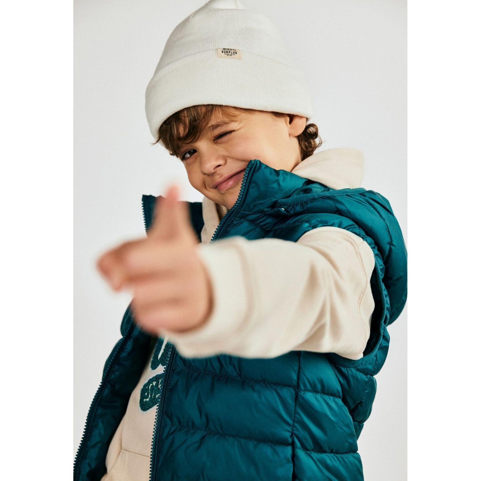 Child wearing cream ribbed beanie with Minoti Surplus label, paired with teal puffer jacket, giving thumbs up gesture against white background