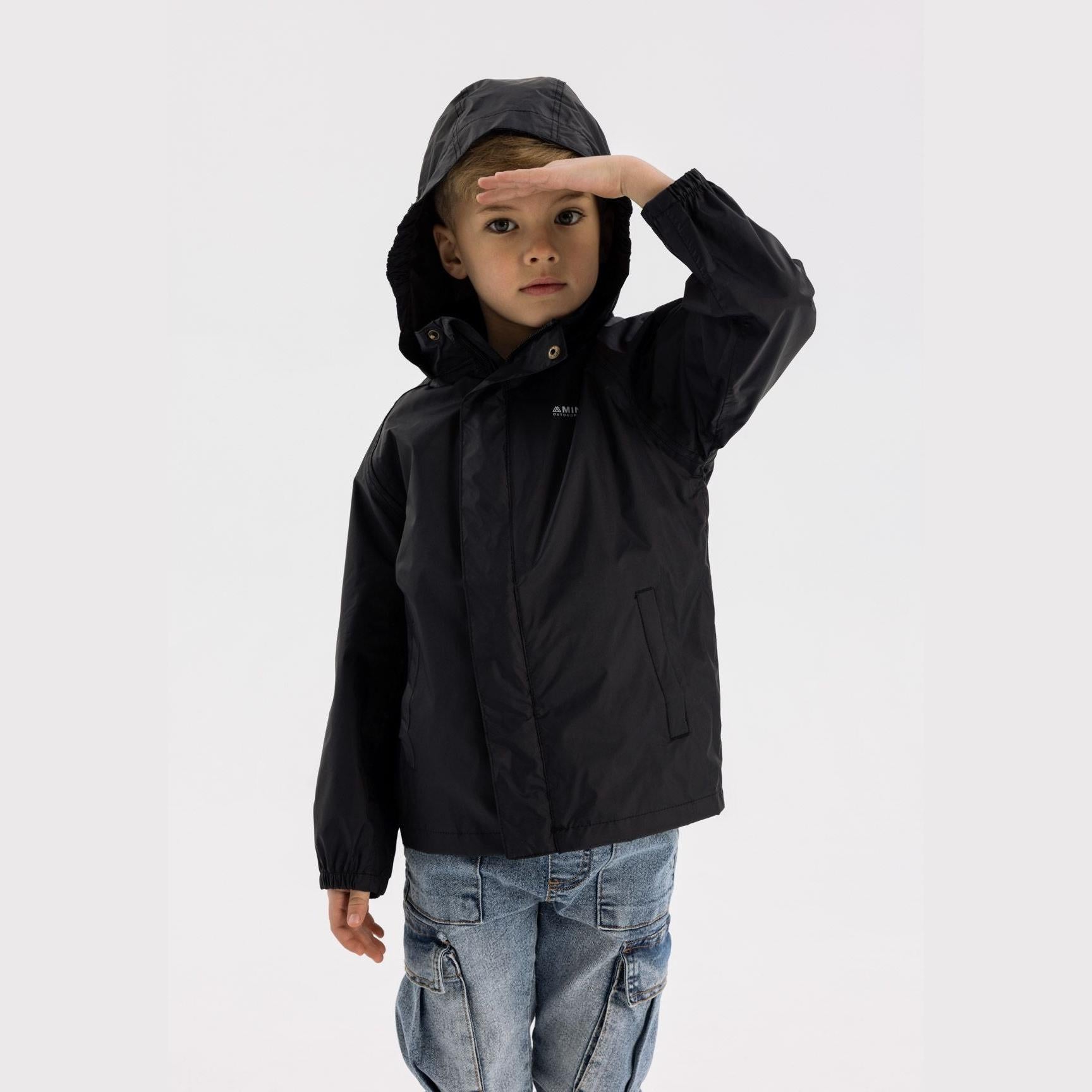Young boy wearing black waterproof jacket with hood up, hand shading eyes, paired with light blue cargo jeans on white background