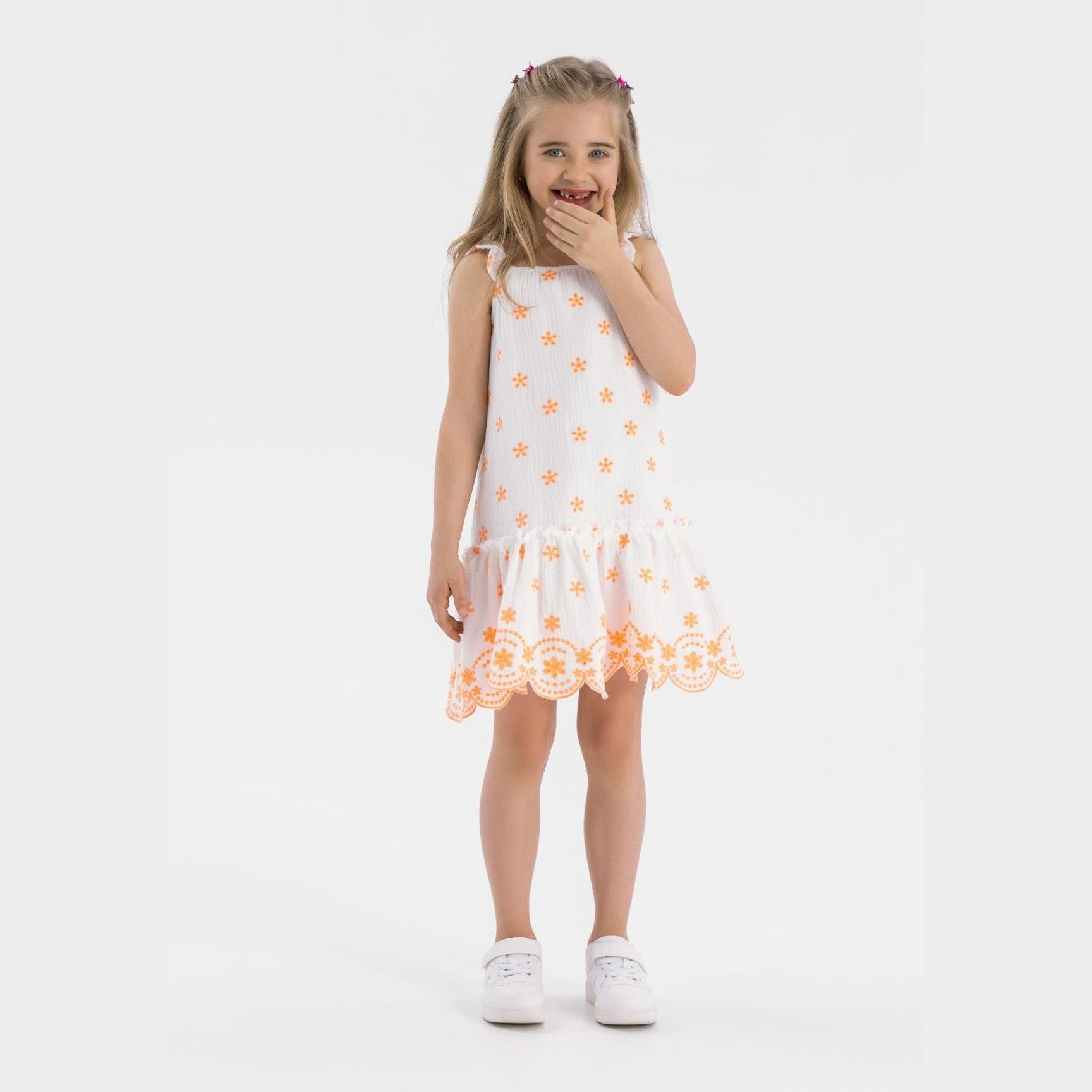 Young blonde girl wearing white cotton muslin dress with orange floral embroidery, flutter straps and scalloped hem, standing against white background