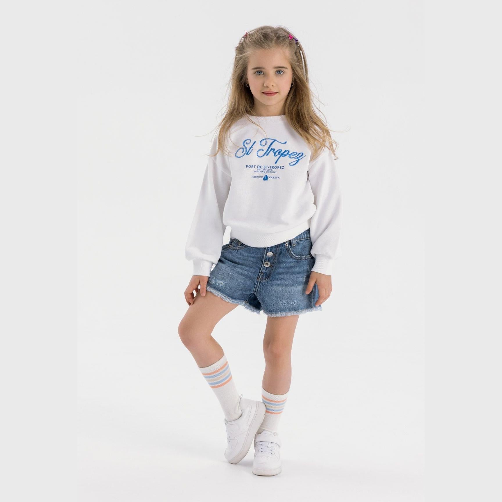 Young girl wearing white fleece sweatshirt with blue St Tropez sailing club print, paired with denim shorts and white sneakers against white background