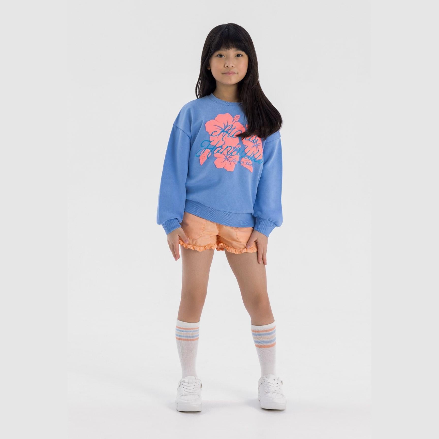 Young girl wearing blue fleece sweatshirt with coral hibiscus print, paired with orange shorts and striped socks, standing against light grey background