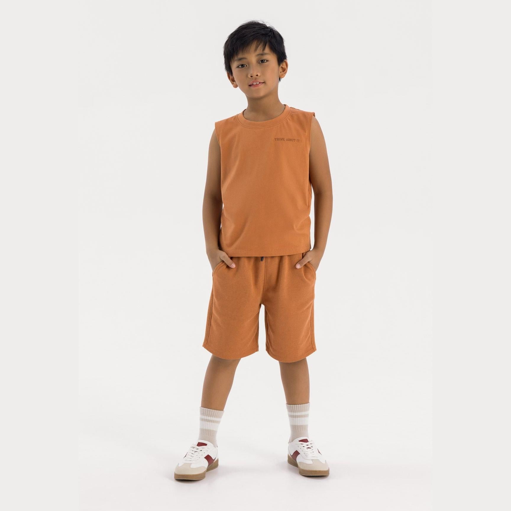 Young boy wearing camel brown vest and shorts set, standing against white background with hands in pockets, wearing white socks and sneakers