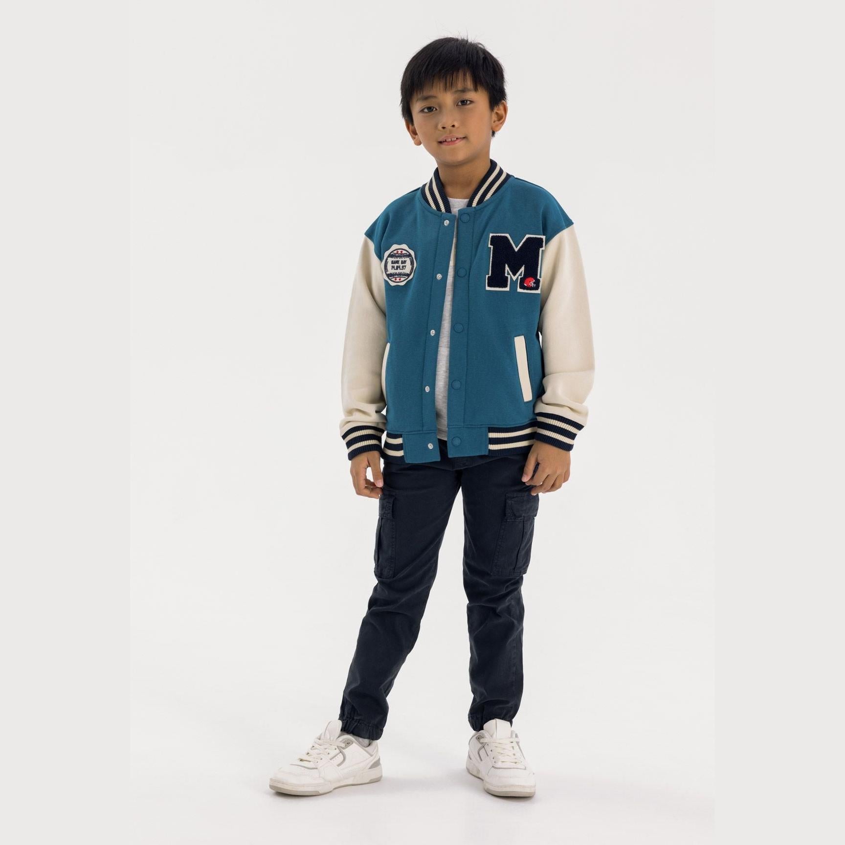 Young boy wearing turquoise and cream fleece baseball jacket with dark jeans and white sneakers against white background