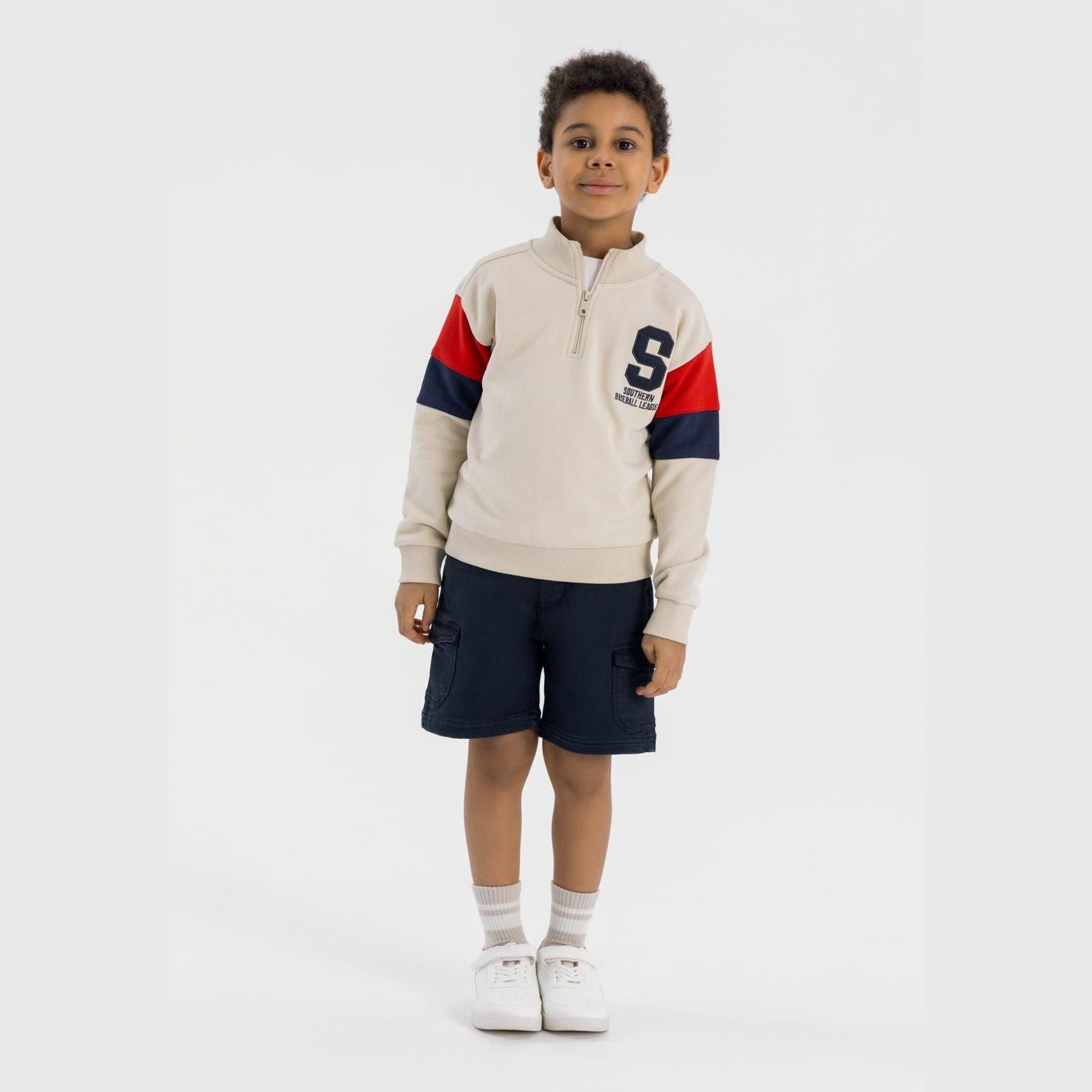 Young boy wearing cream half-zip sweatshirt with red and navy sleeve details, paired with navy shorts, full body shot on white background