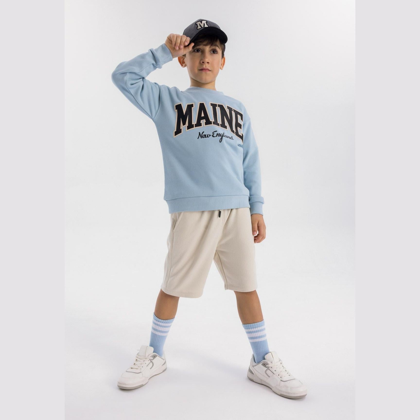 Child model wearing light blue fleece sweatshirt with Maine New England print, paired with beige shorts and striped socks, against white studio background