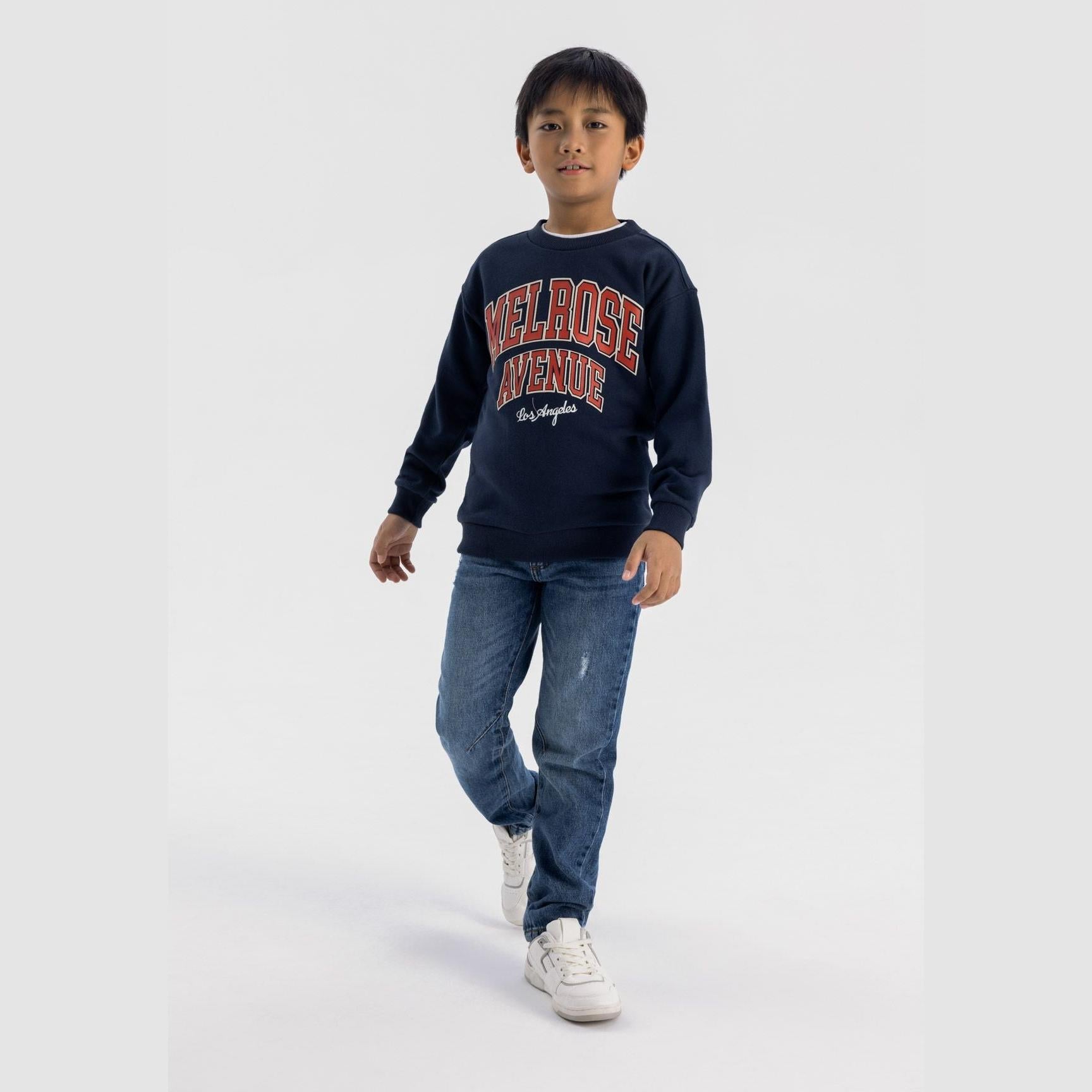 Boy wearing navy fleece sweatshirt with Melrose Avenue Los Angeles print, paired with distressed jeans and white sneakers, walking pose on white background