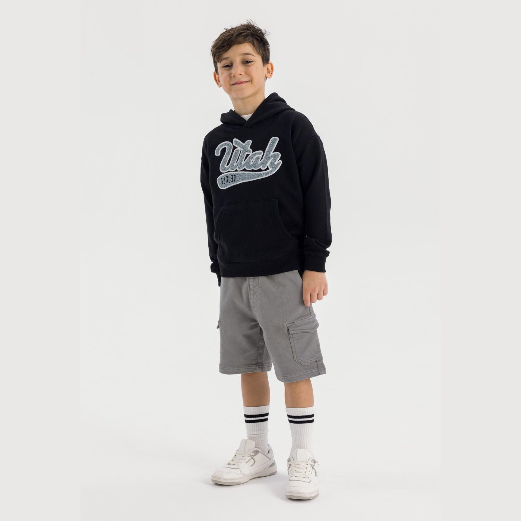 Boy wearing black oversized hoodie with grey embroidered Wtak logo, paired with grey cargo shorts and white sneakers, standing against light grey background