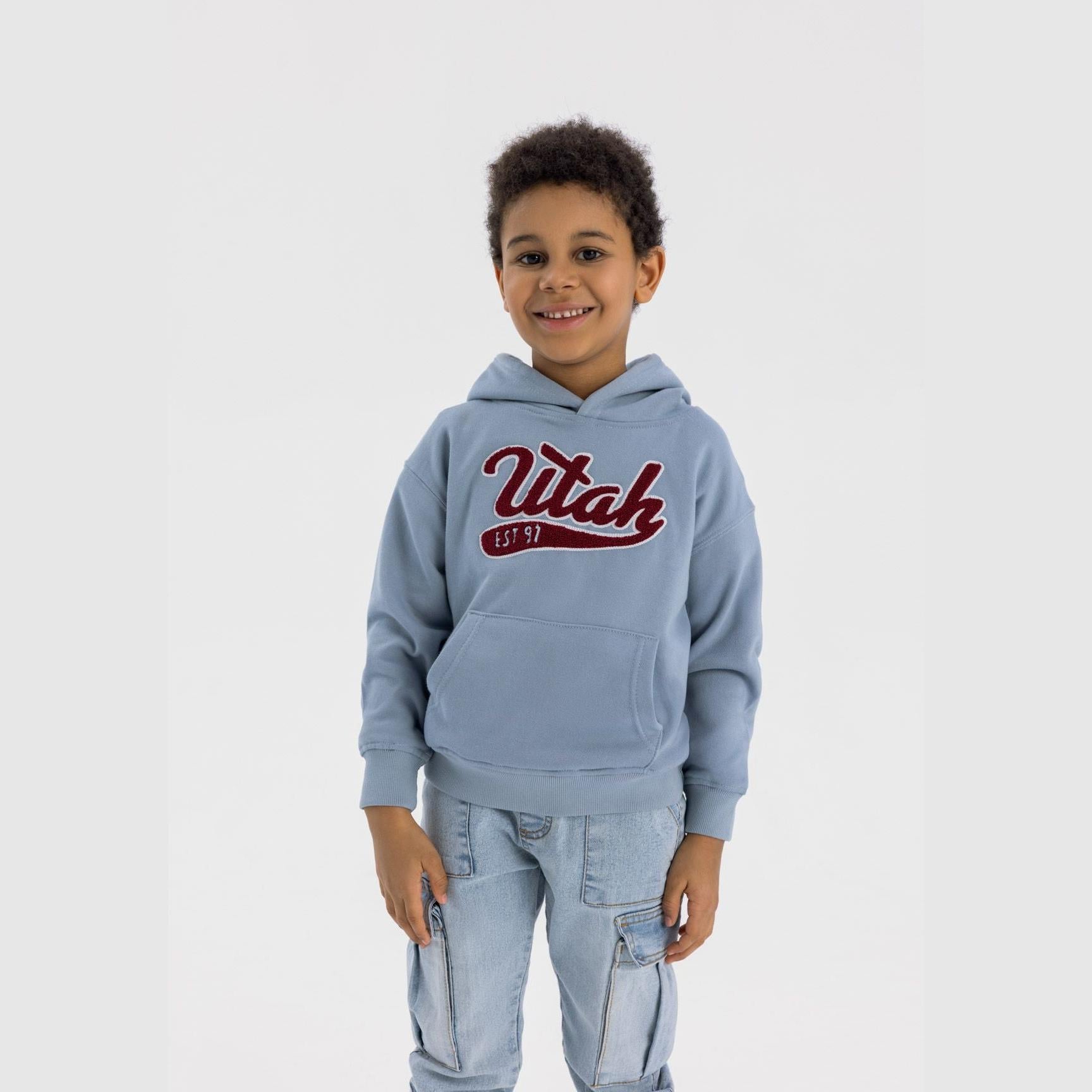 Young boy wearing light blue oversized hoodie with 'Utah EST 97' embroidered logo, paired with light blue jeans, standing against white background