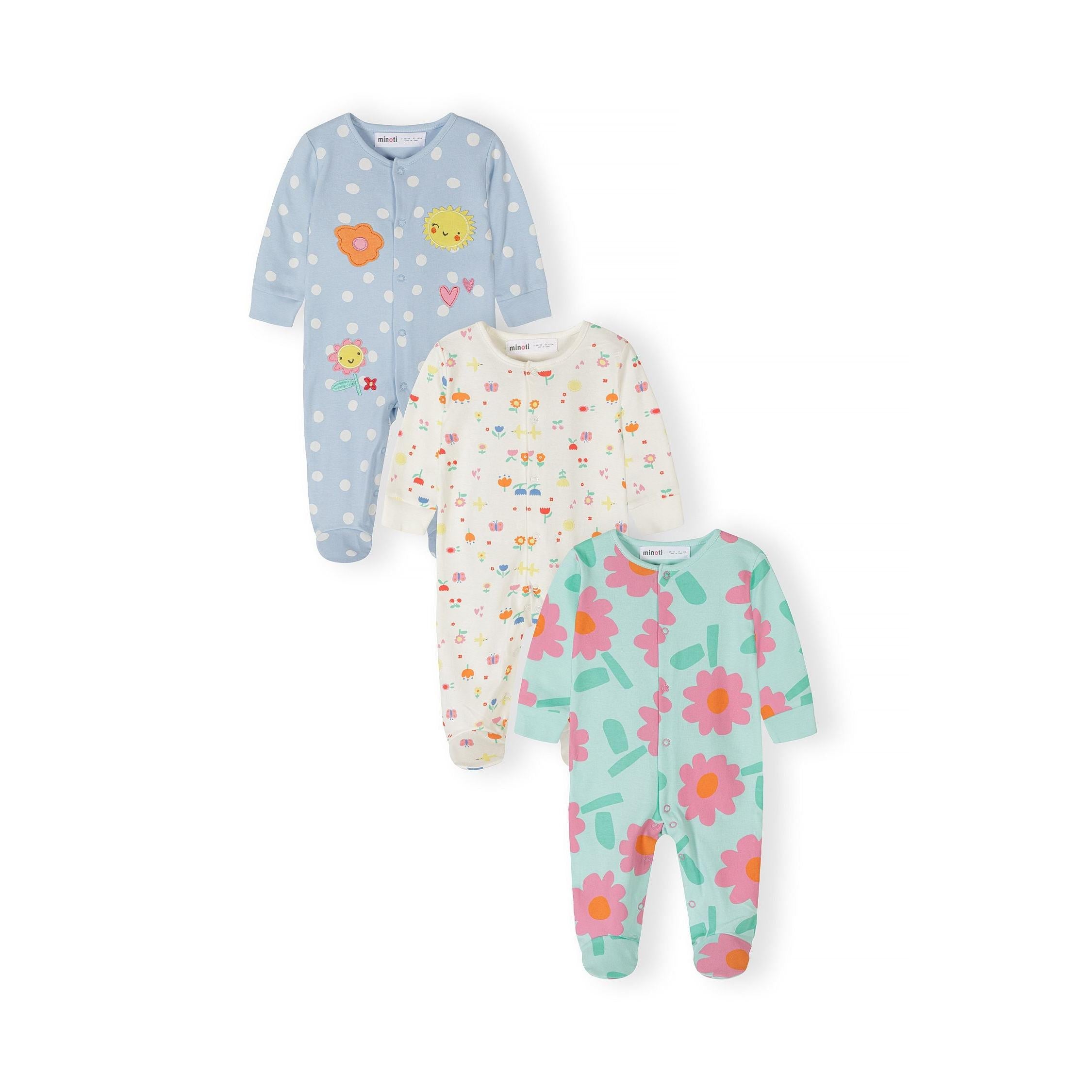 Three baby sleepsuits laid flat showing light blue polka dot with sun and flower appliqués, cream with scattered colorful prints, and mint green with pink floral pattern