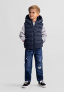 Young boy wearing navy blue padded hooded gilet with zip pockets and casual jeans, standing confidently against gray background