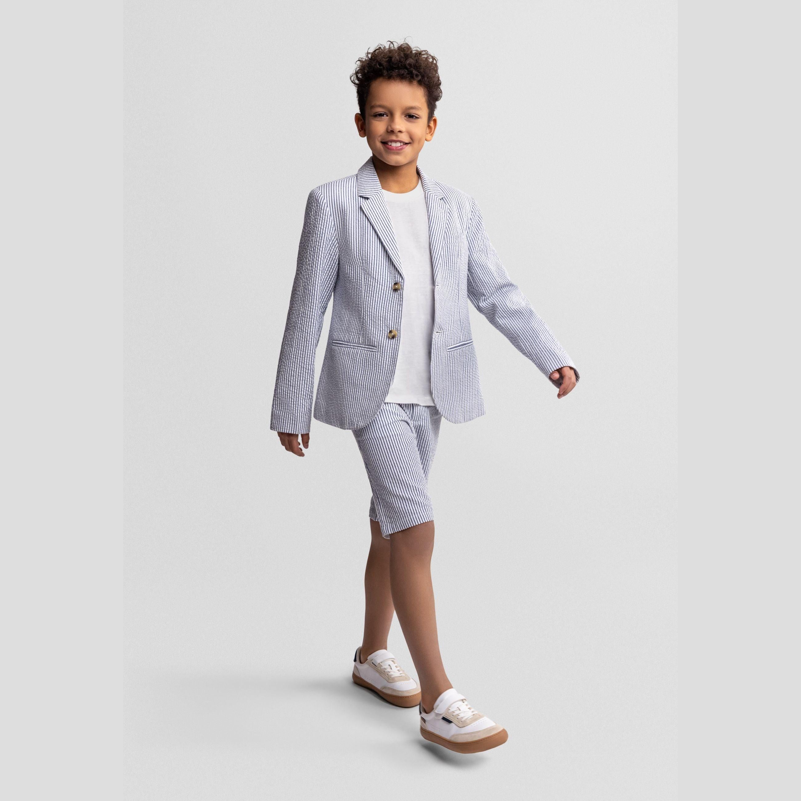 Young boy wearing navy striped cotton blazer with white t-shirt and matching shorts, standing confidently against light grey background