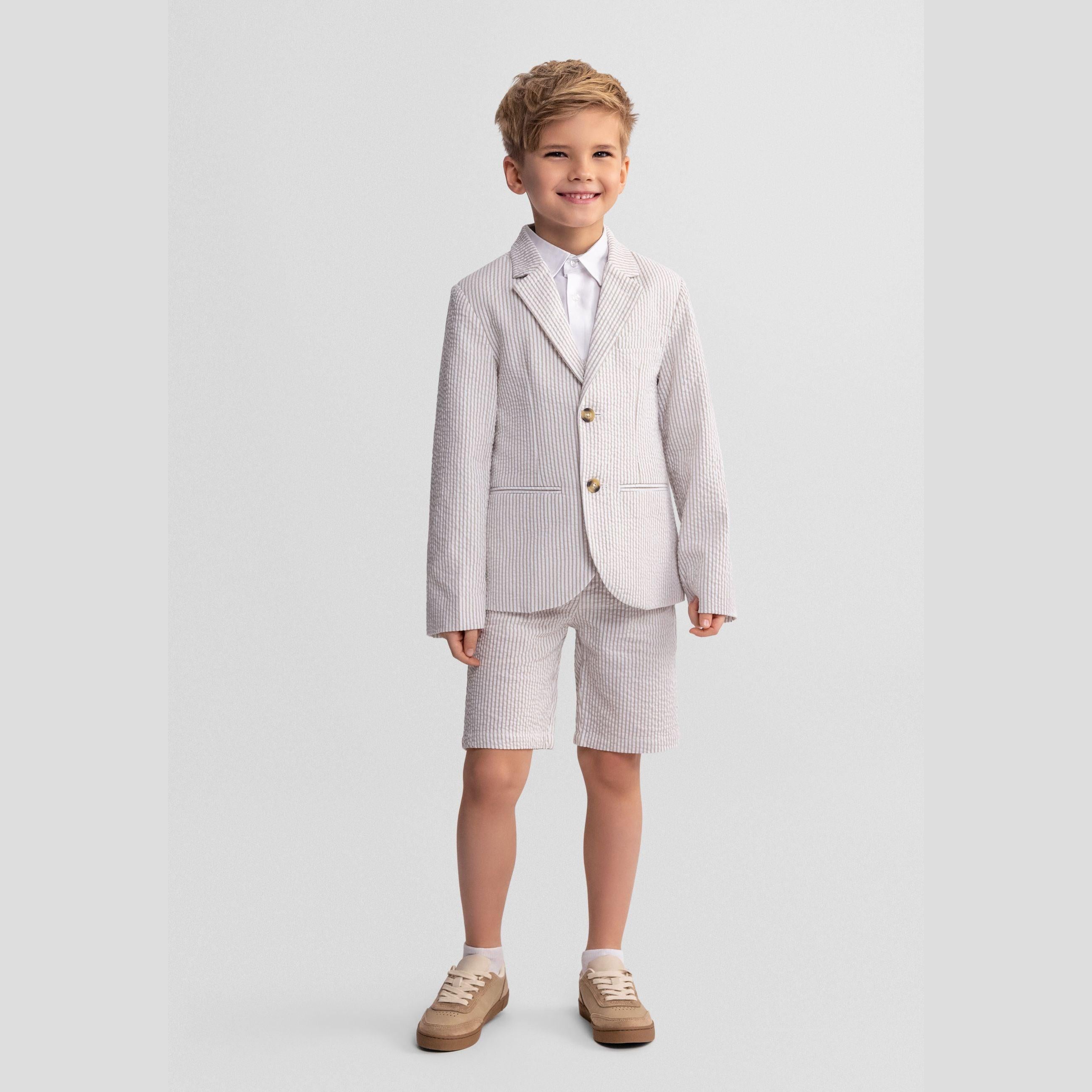 Young boy wearing stone stripe tailored cotton suit shorts with matching blazer and white shirt, standing against light grey background