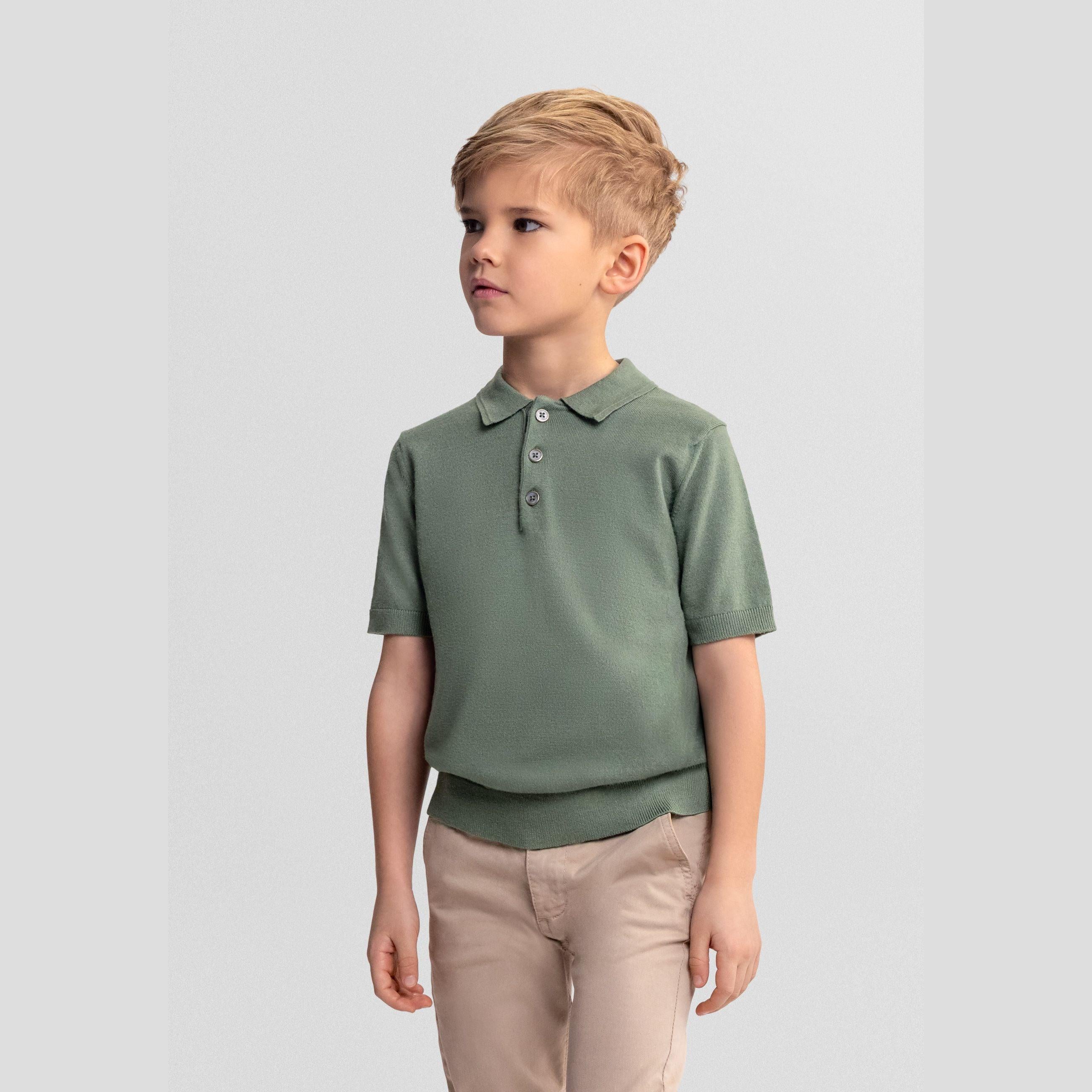 Young boy wearing forest green knitted polo shirt with beige trousers, standing against light grey background, looking to the side