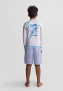 Boy wearing a long sleeve rash vest with blue surf design and light blue shorts, standing barefoot facing away.
