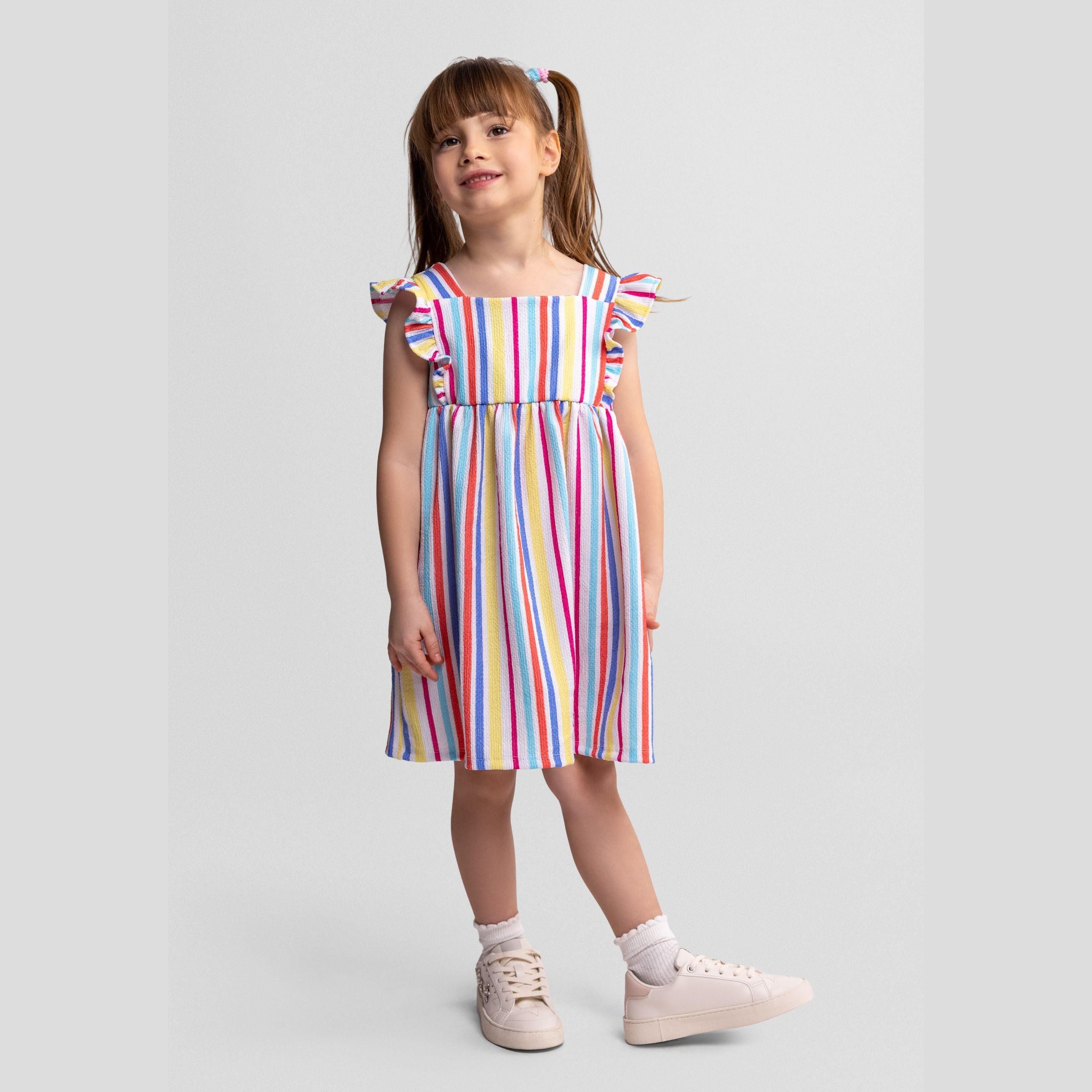Young girl wearing multicolour striped sleeveless dress with flutter sleeves, standing against white background with hands at sides, smiling at camera