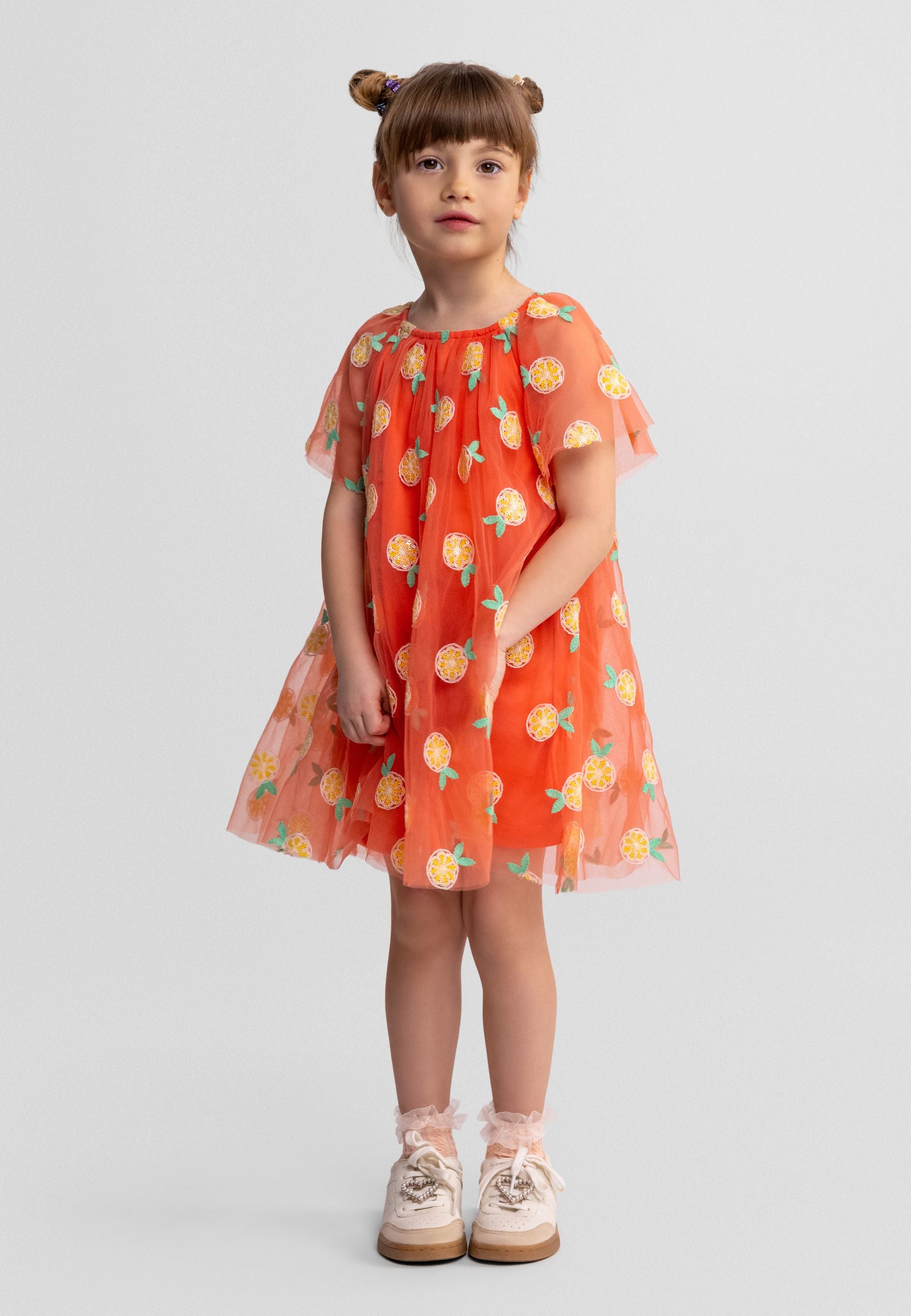 Young girl wearing orange tulle dress with sequin angel sleeves and citrus print, standing against white studio background with hands at sides