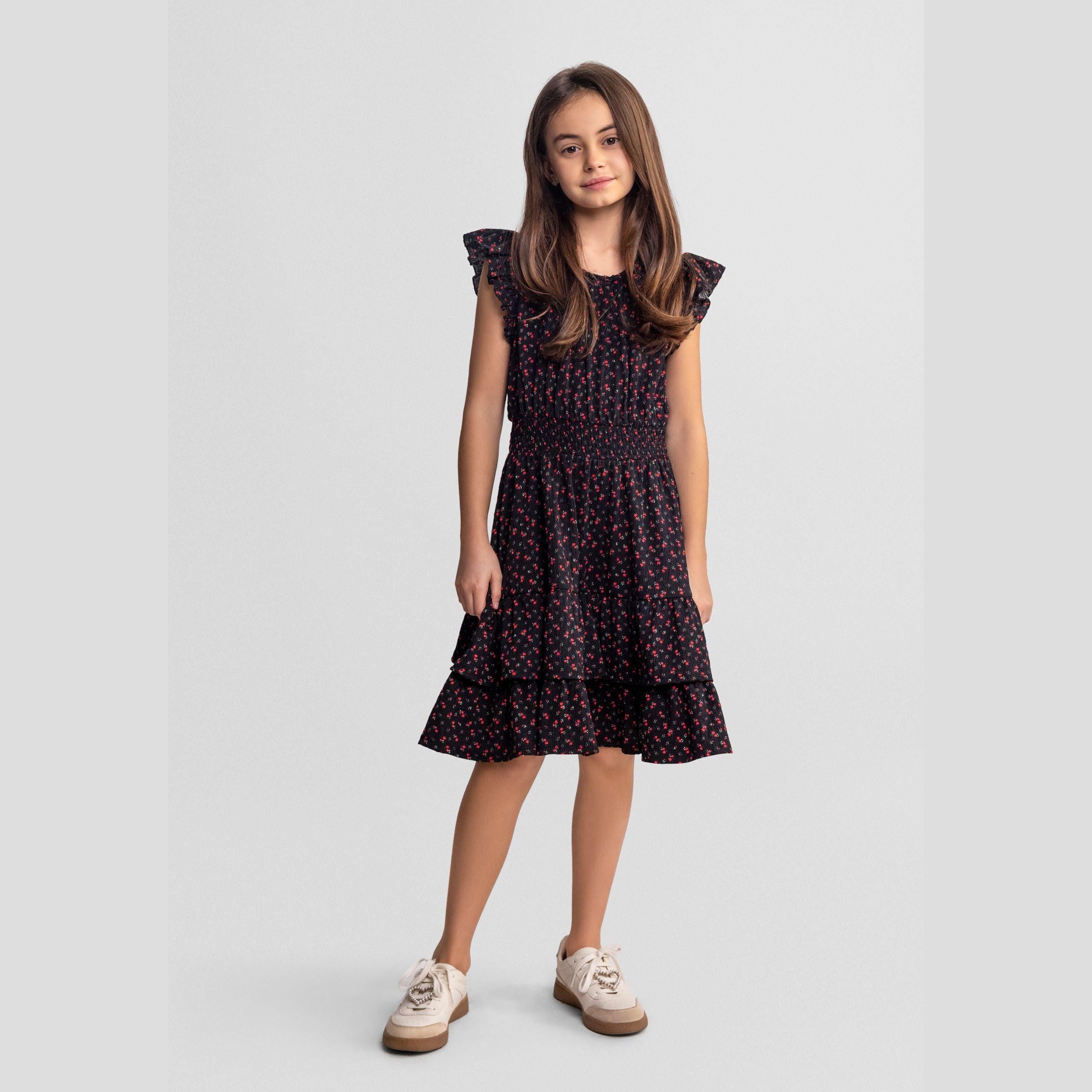 Young girl wearing black floral A-line dress with red flower print and frill sleeves, standing against light grey background in casual pose