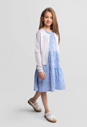 Young girl wearing a white cardigan with wavy trim over a blue floral dress, smiling with casual shoes on gray background.