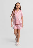 Young girl wearing a pink satin striped button-front pyjama set with shorts and soft slippers, showcasing a stylish design.