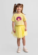 Smiling young girl wearing a pale yellow t-shirt with rainbow design and a matching yellow skirt standing on a gray background