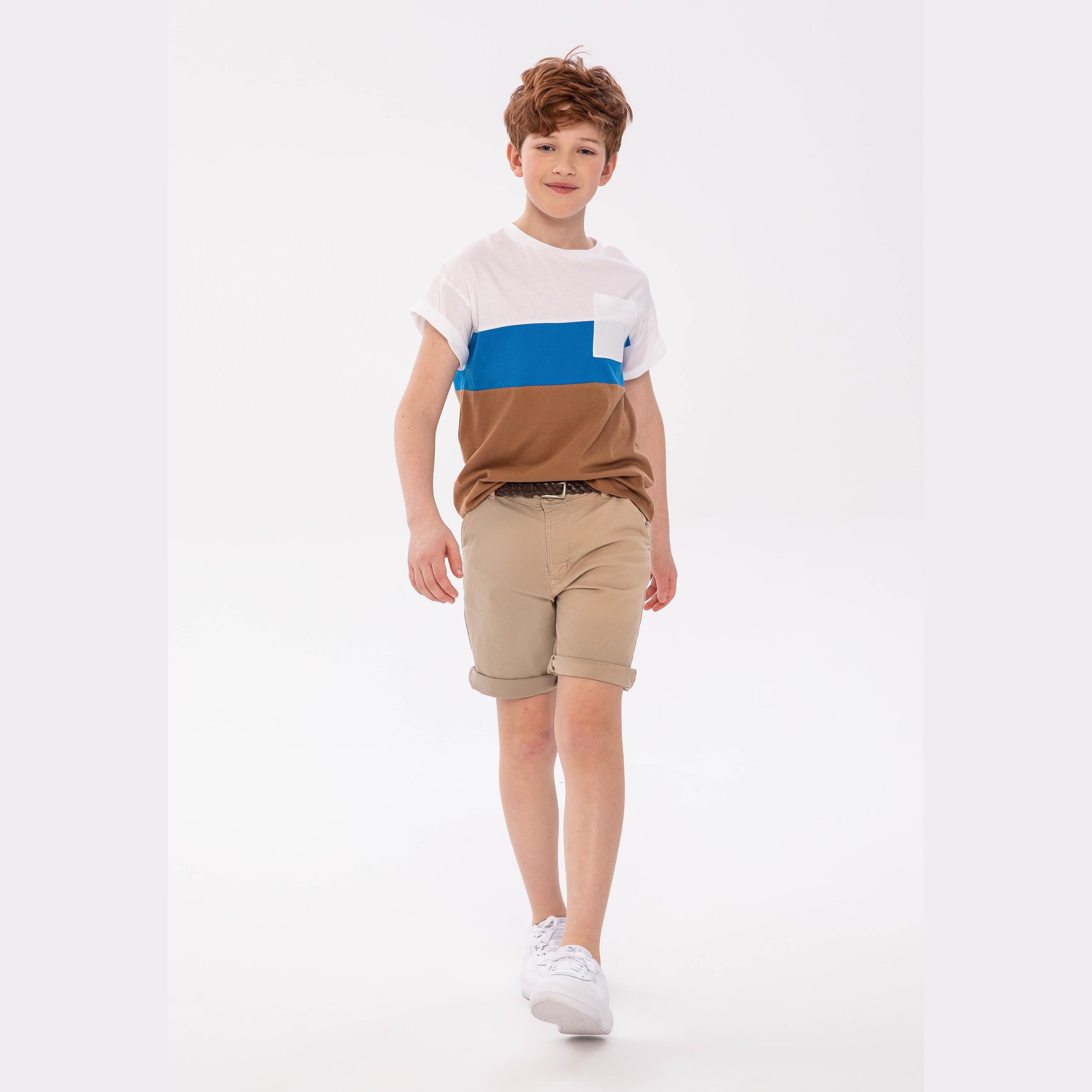 Boy wearing white t-shirt with blue and brown contrast panels, paired with beige shorts, walking against white studio background