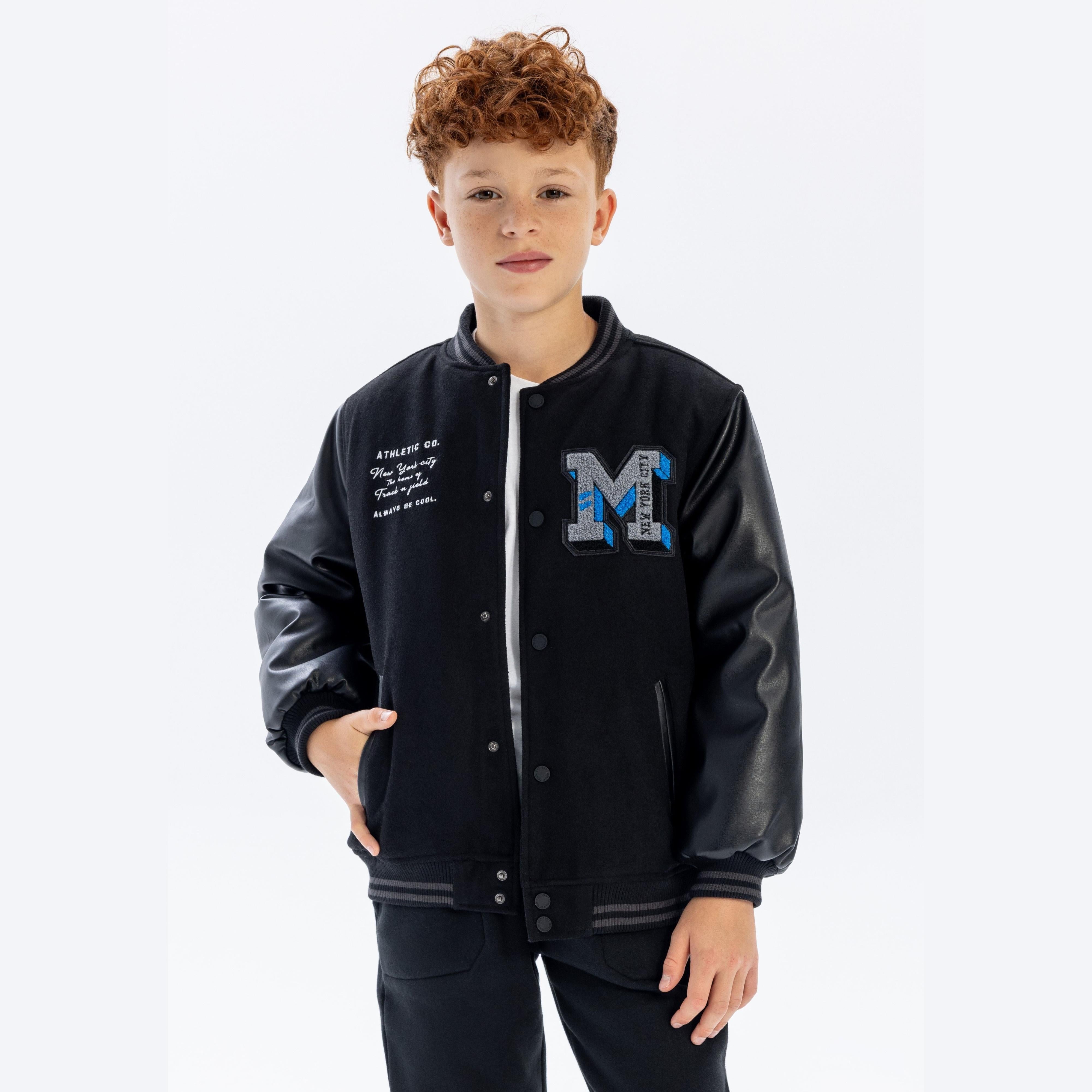 Boy wearing black baseball jacket with faux leather sleeves, featuring large M logo and Athletic Co. embroidery, standing against white background