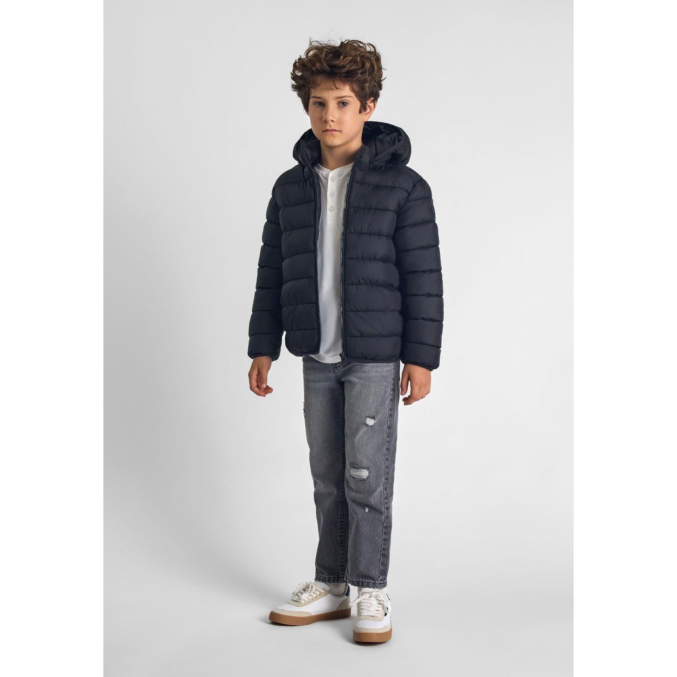 Boy wearing black padded jacket with hood over light grey t-shirt and distressed jeans, standing against light grey background