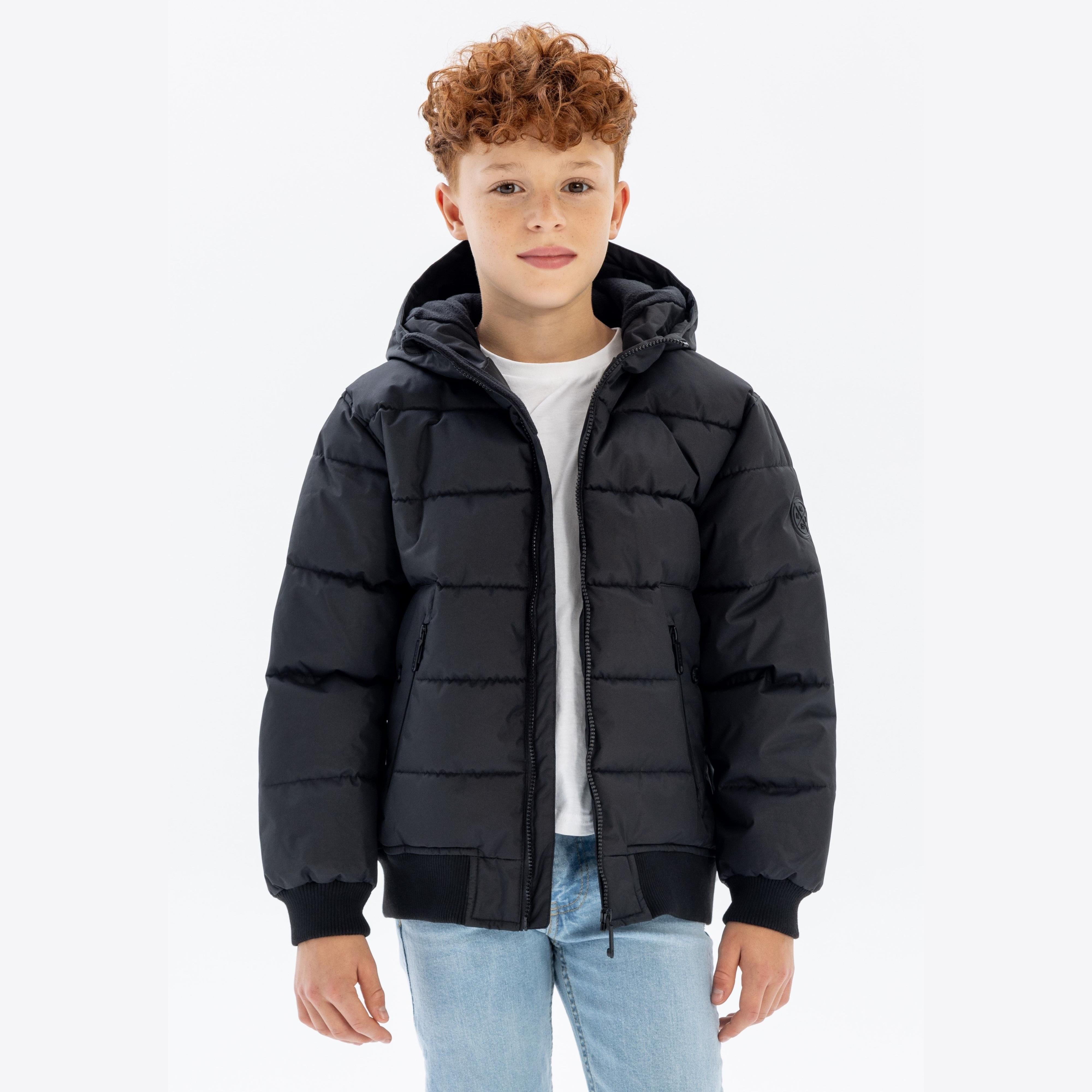 Boy with curly hair wearing black hooded padded jacket with white t-shirt and jeans, standing against white background