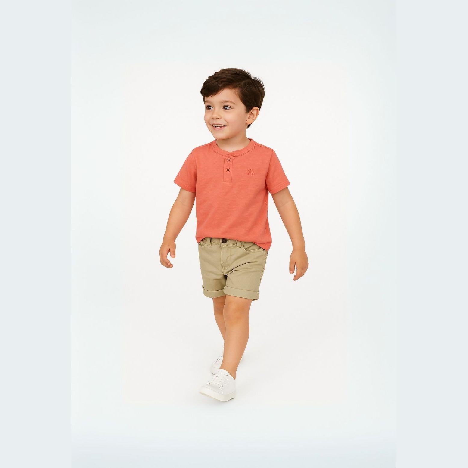 Young boy wearing coral henley t-shirt with button neck and beige shorts, walking against white background