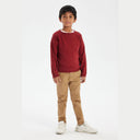 Young boy wearing a red knitted crew neck jumper with a v-insert, paired with tan trousers and white sneakers.
