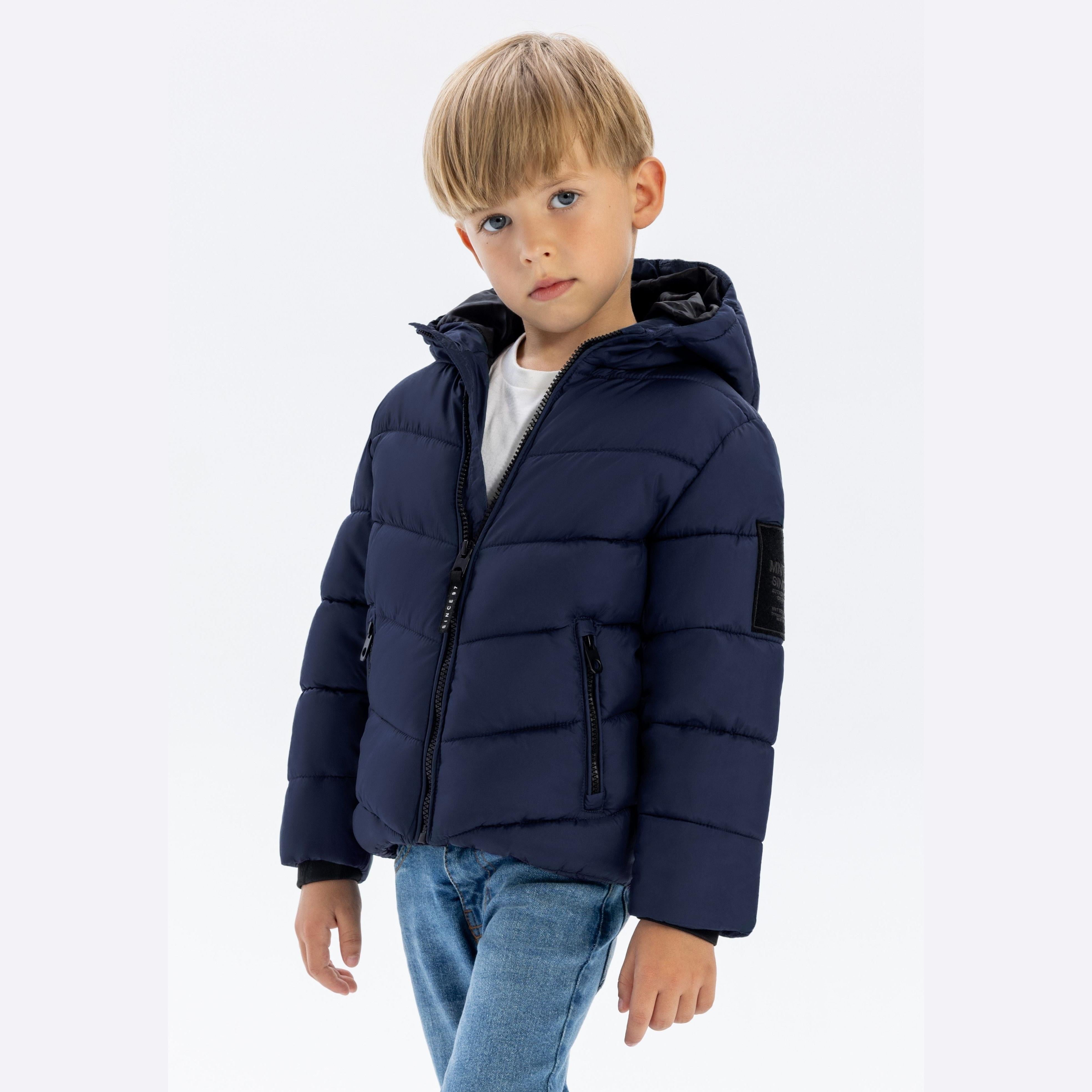 Young blonde boy wearing navy quilted hooded jacket with reflective patch detail, paired with blue jeans, standing against white background