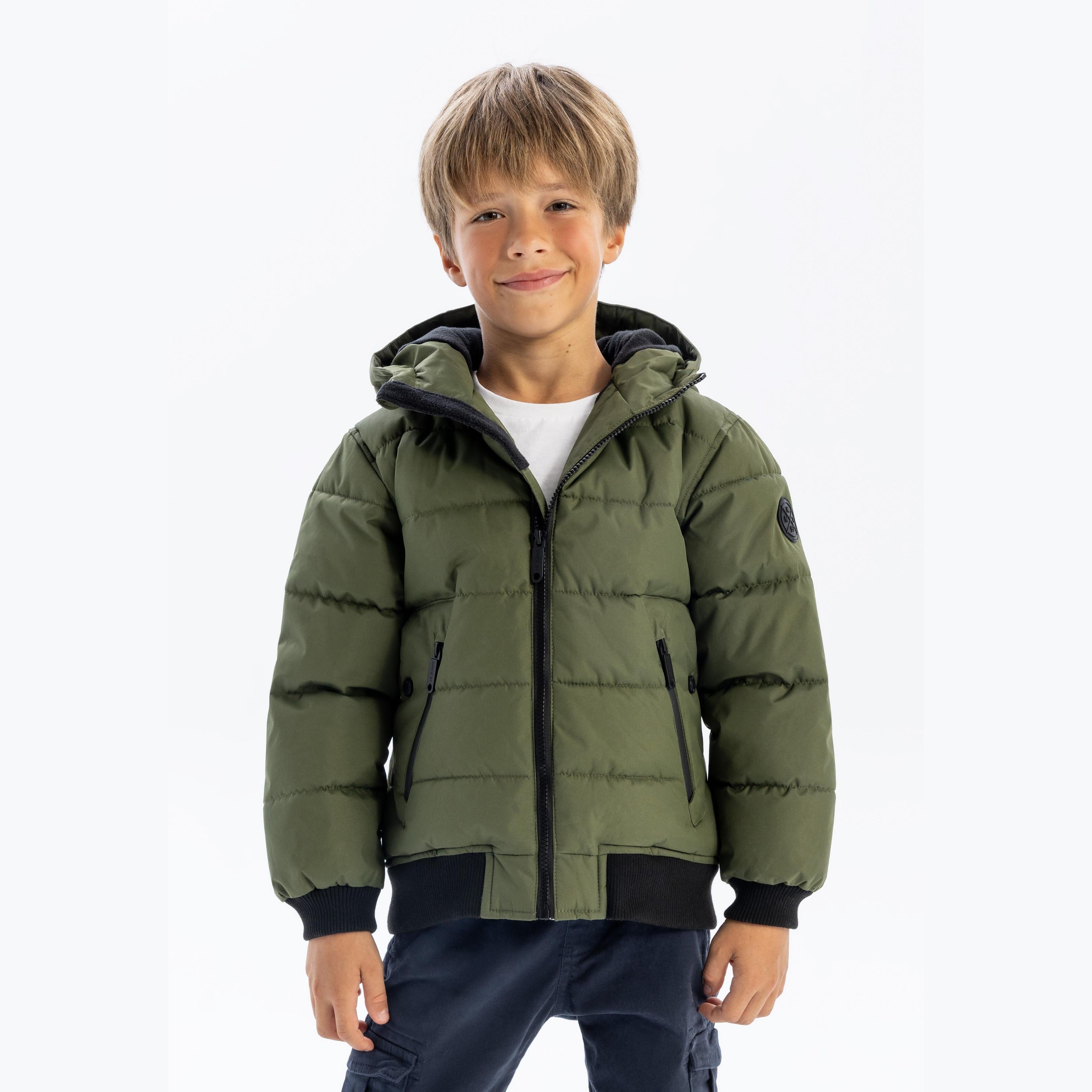 Young boy wearing khaki padded jacket with hood, white t-shirt and navy trousers, smiling at camera against white background