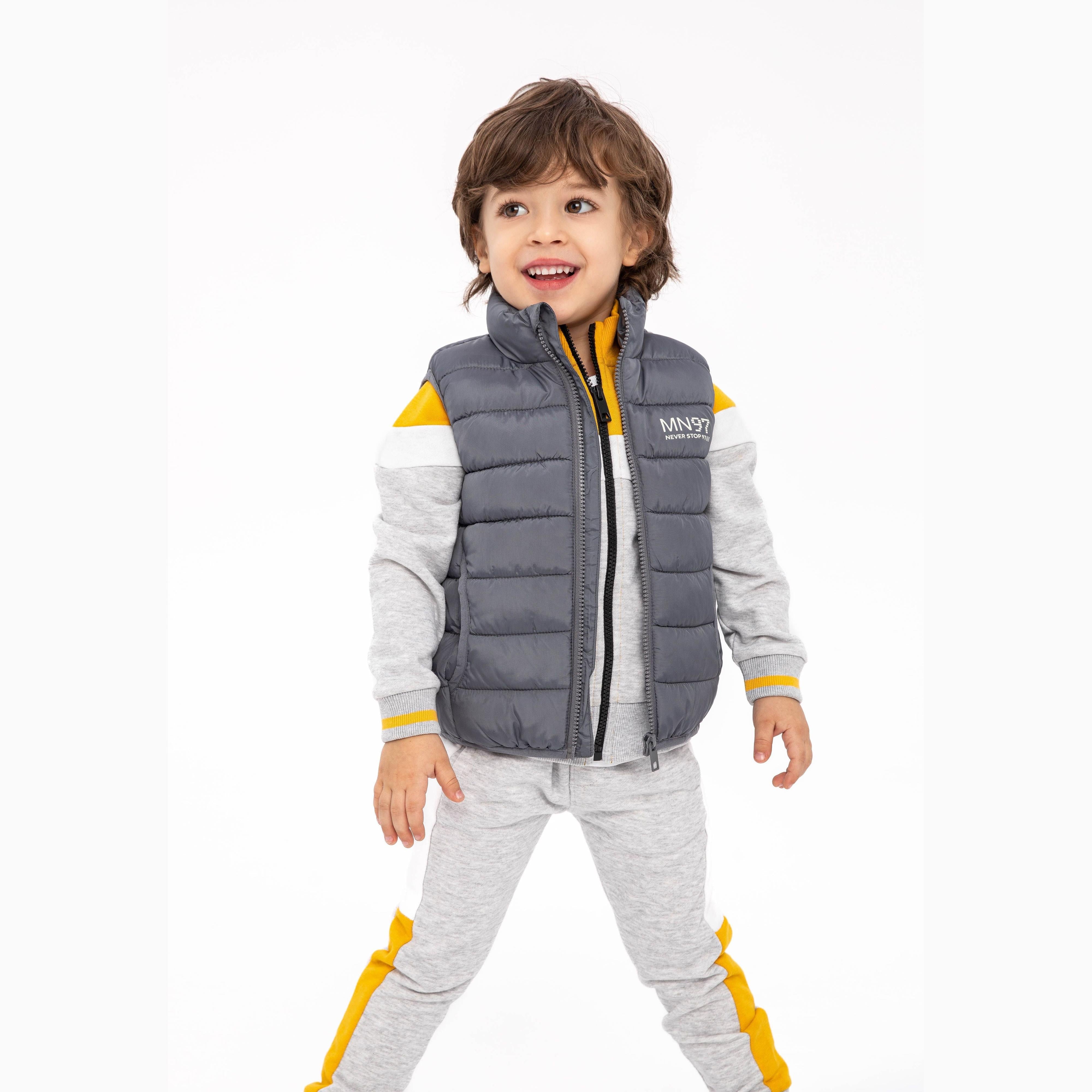 Young boy wearing grey padded gilet over grey tracksuit with yellow trim, smiling on white background