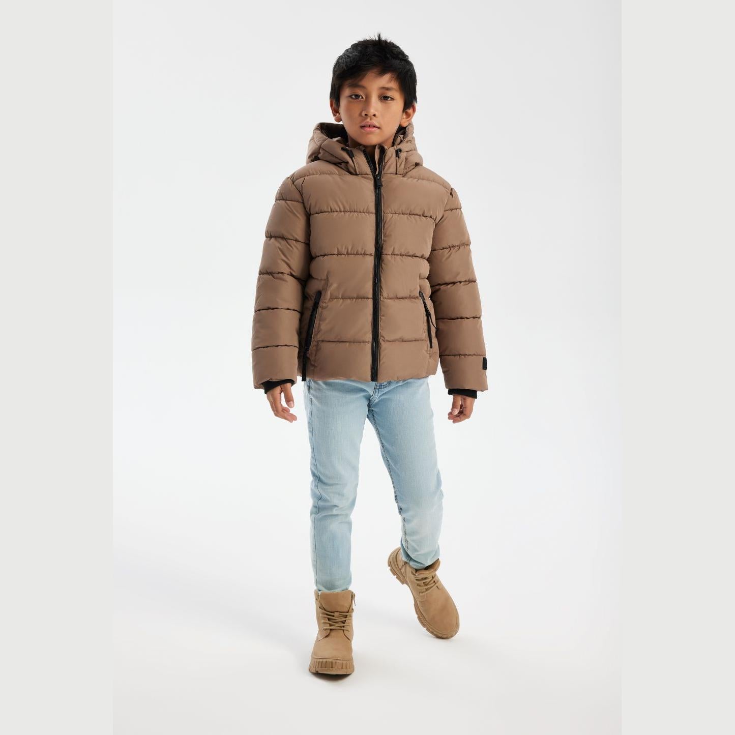 Young boy wearing beige padded jacket with hood and light blue jeans, standing against white background