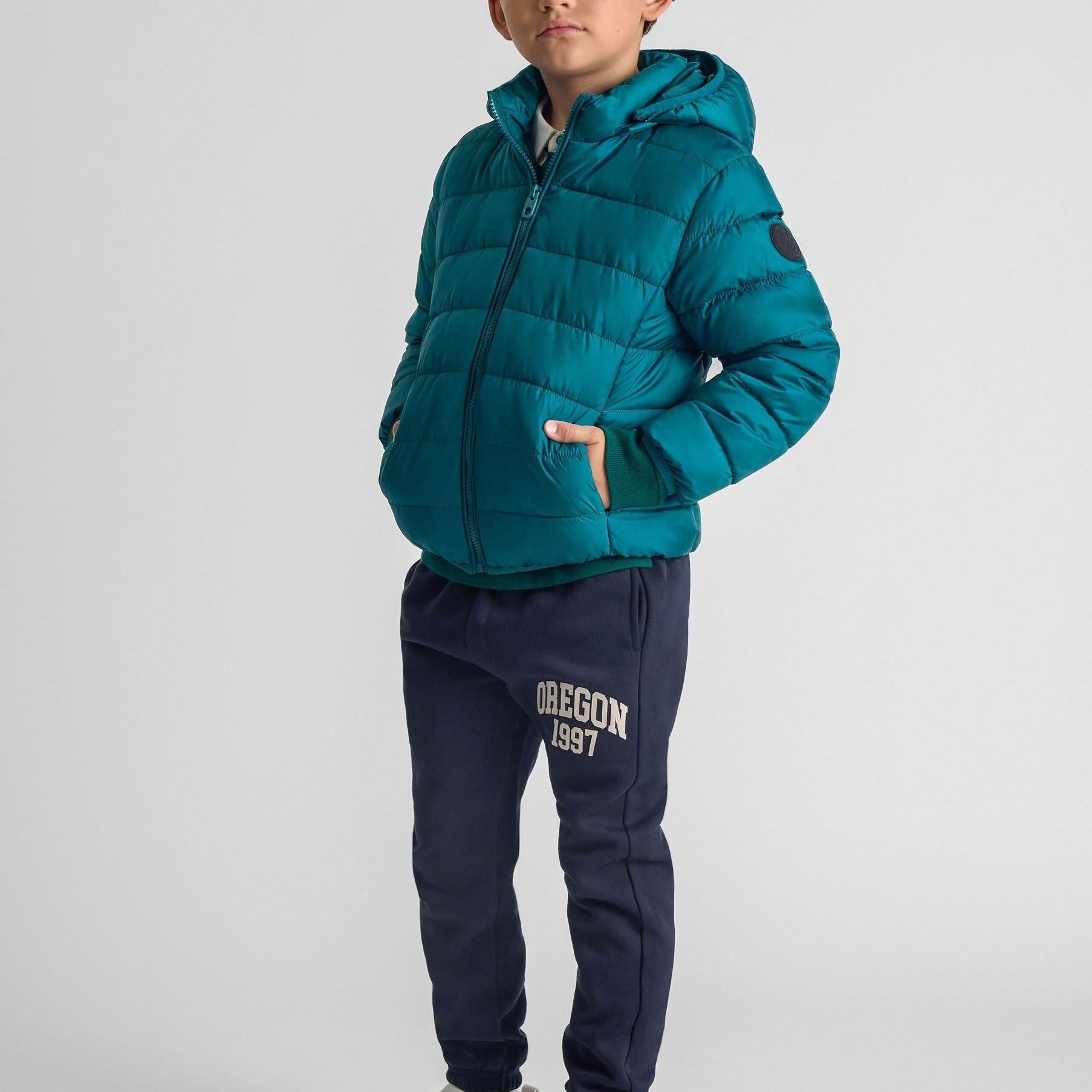 Child wearing turquoise padded jacket with detachable hood and navy joggers with Oregon 1997 text, standing against light grey background