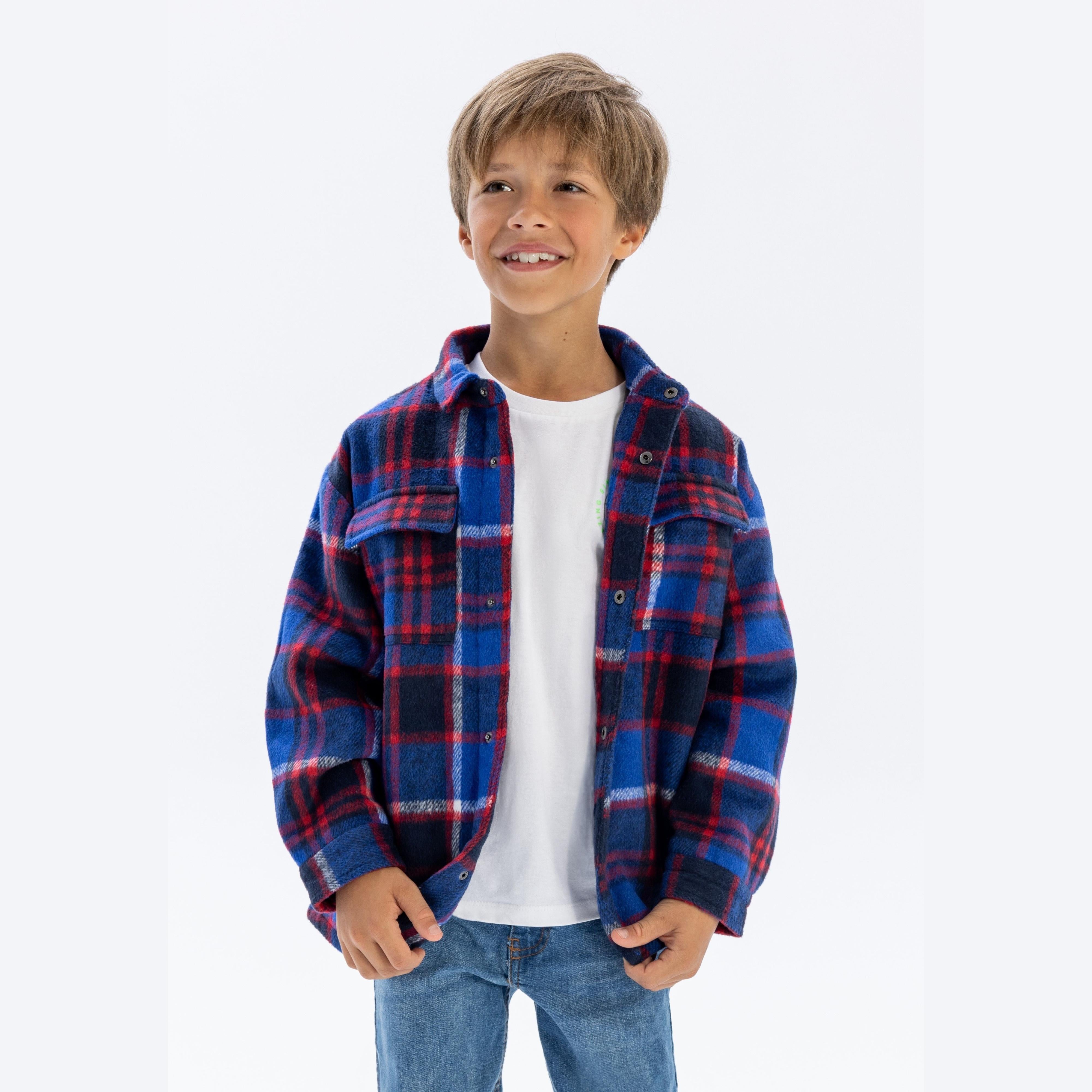 Young boy wearing blue check padded shirt jacket over white t-shirt and jeans, smiling against white studio background