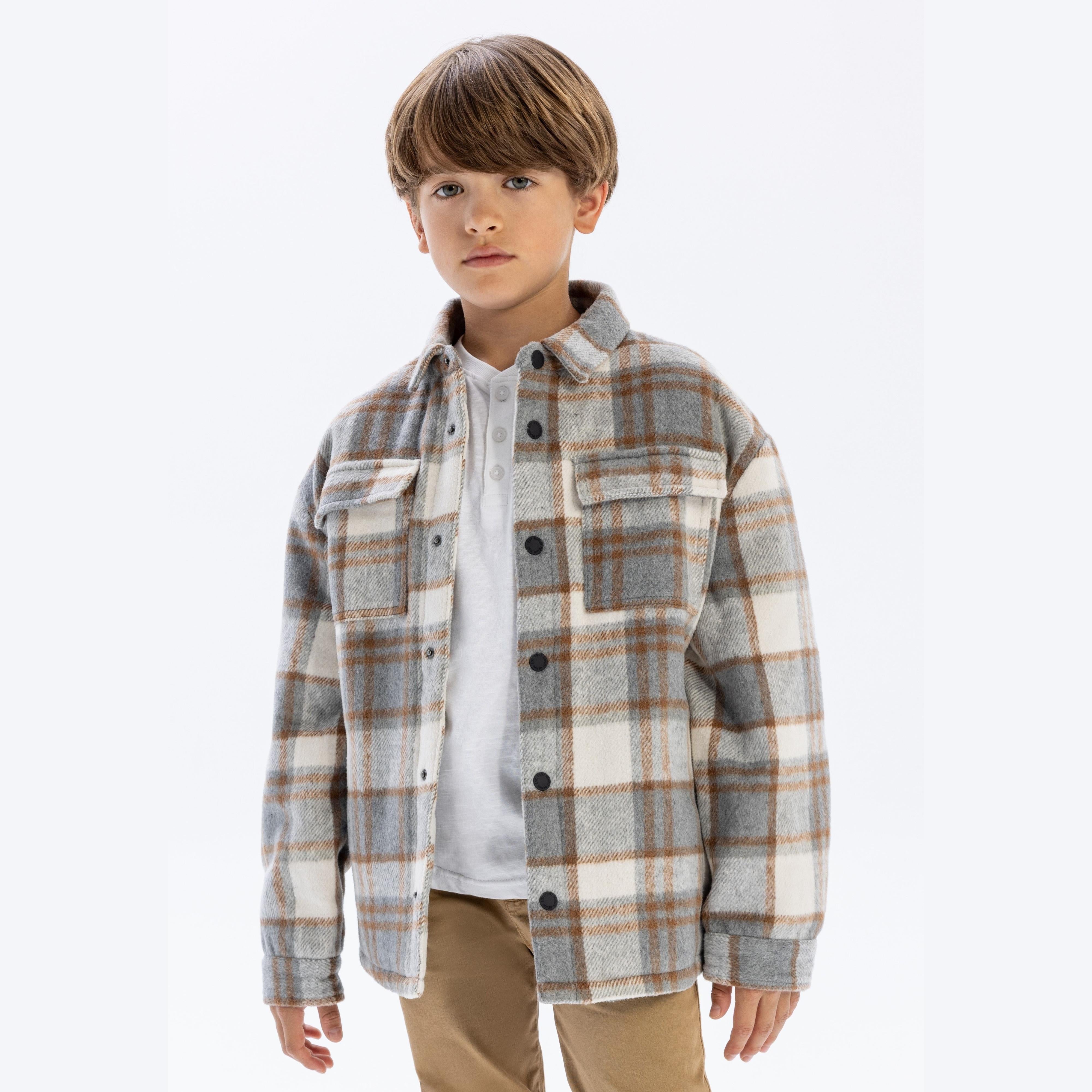 Young boy wearing cream check padded shirt jacket over white top and beige trousers, standing against white background