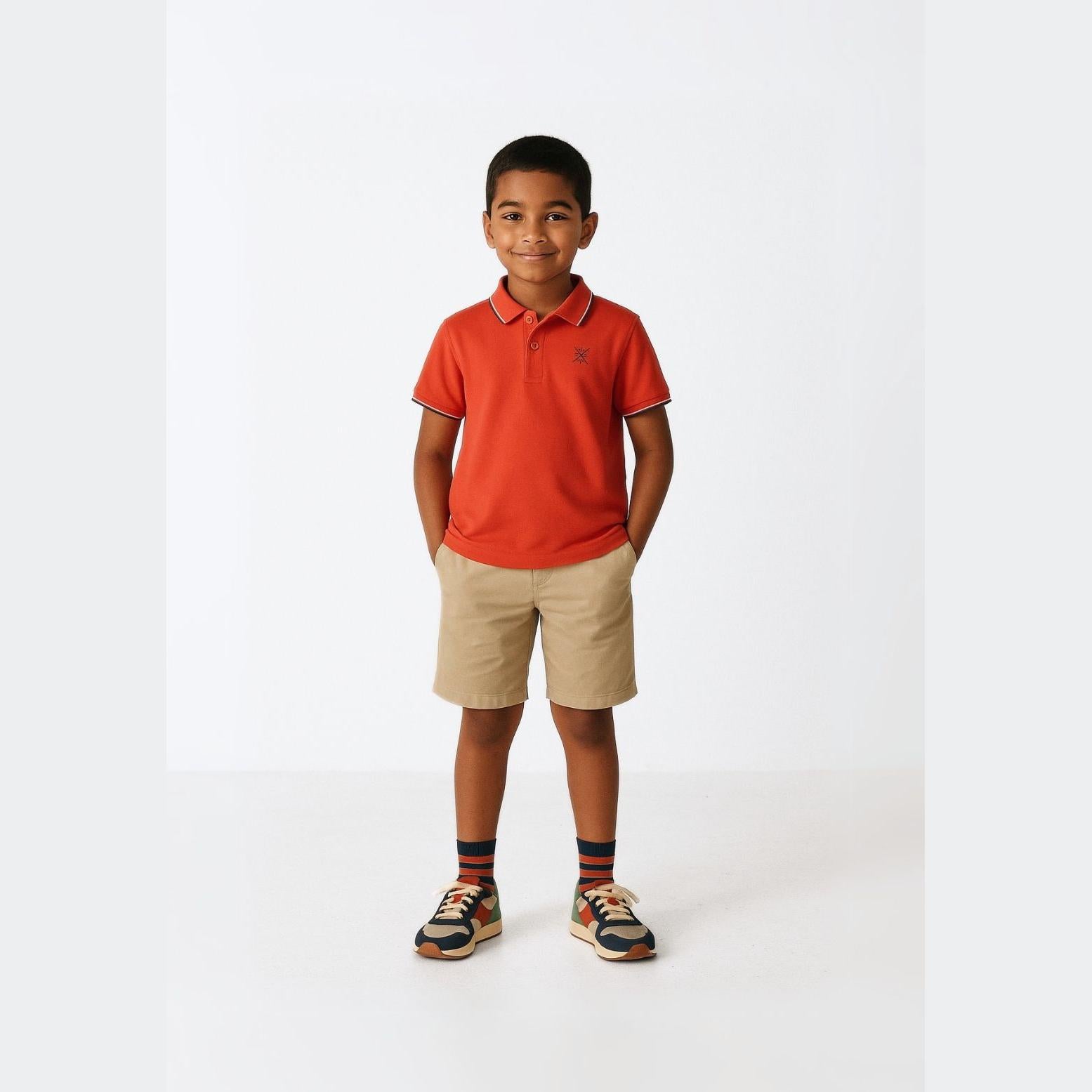 Young boy wearing red polo shirt with contrast tipping and beige shorts, standing against white background with hands in pockets